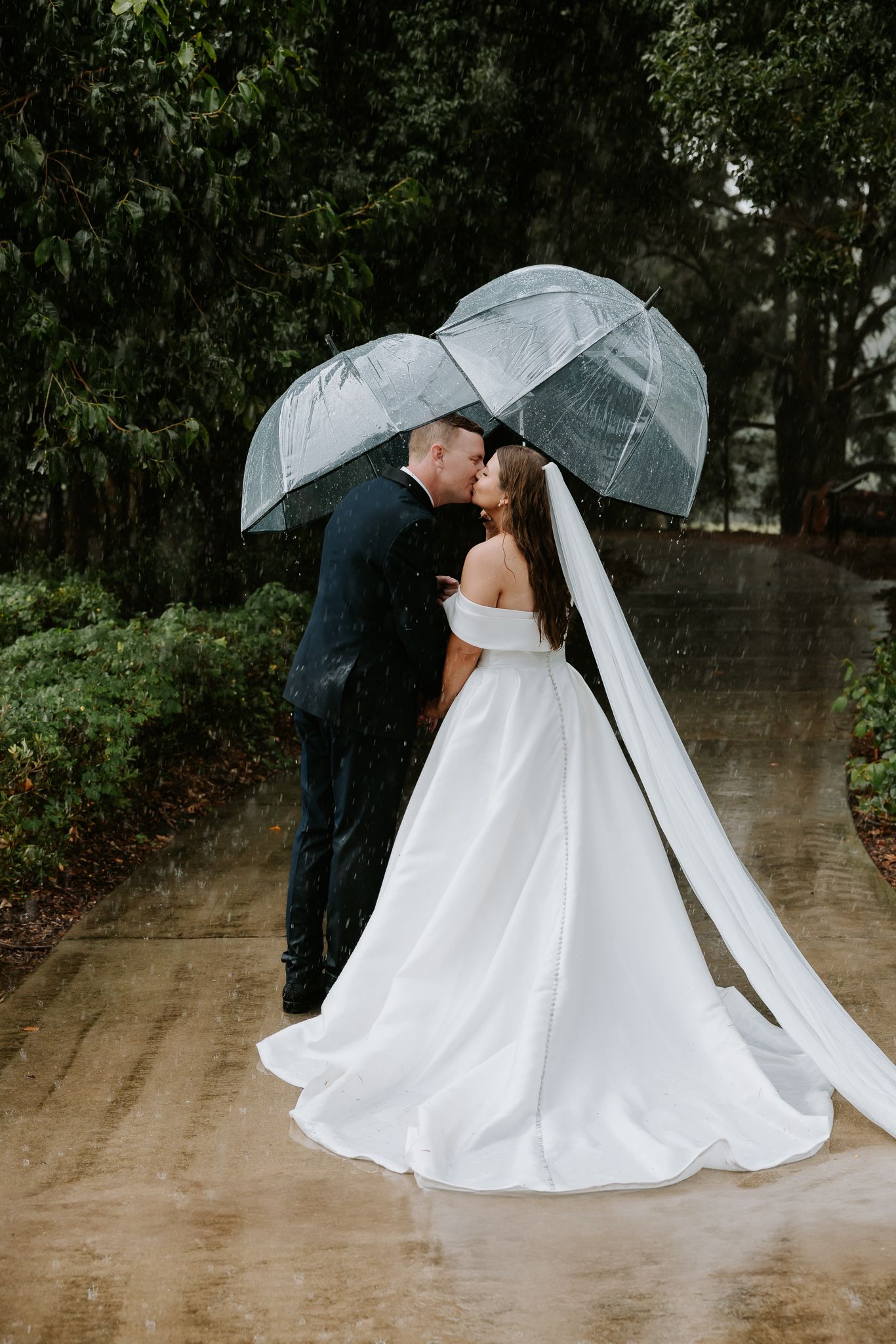Romantic rainy wedding photos under clear umbrella with flowing white dress.