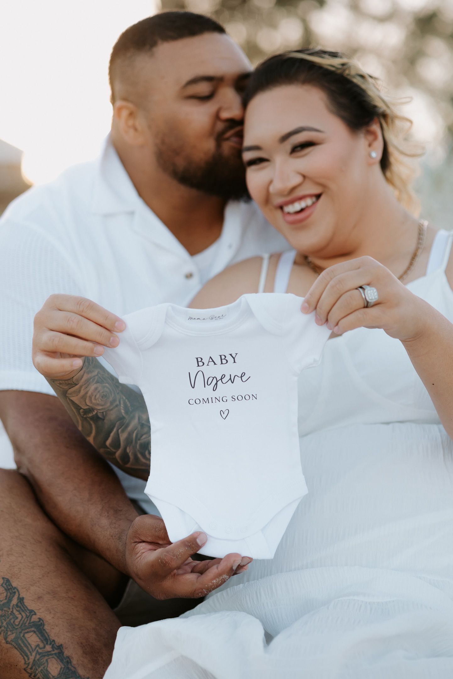 Couple holds up white baby onesie with text 'Baby Coming Soon' during pregnancy announcement photoshoot at sunset.
