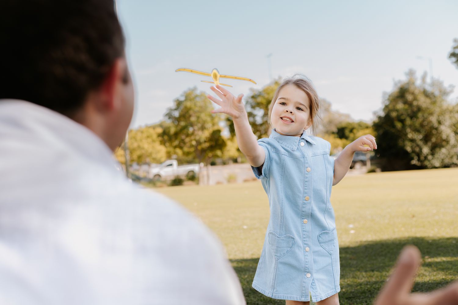 A happy child plays with a toy airplane outdoors with an adult on a sunny day in a green park.