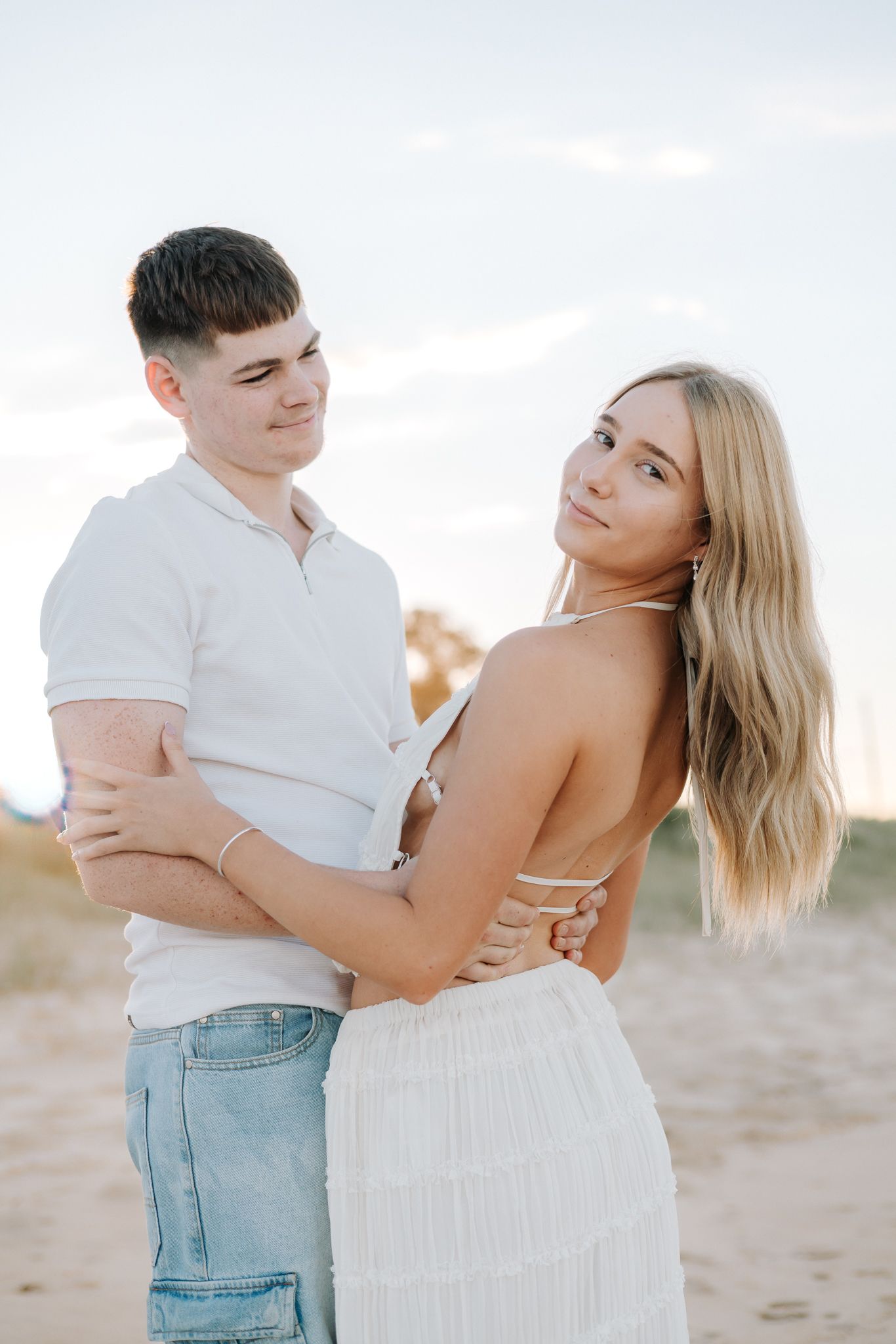 A young couple embraces on a sandy beach at sunset, woman in white dress looks back over shoulder.