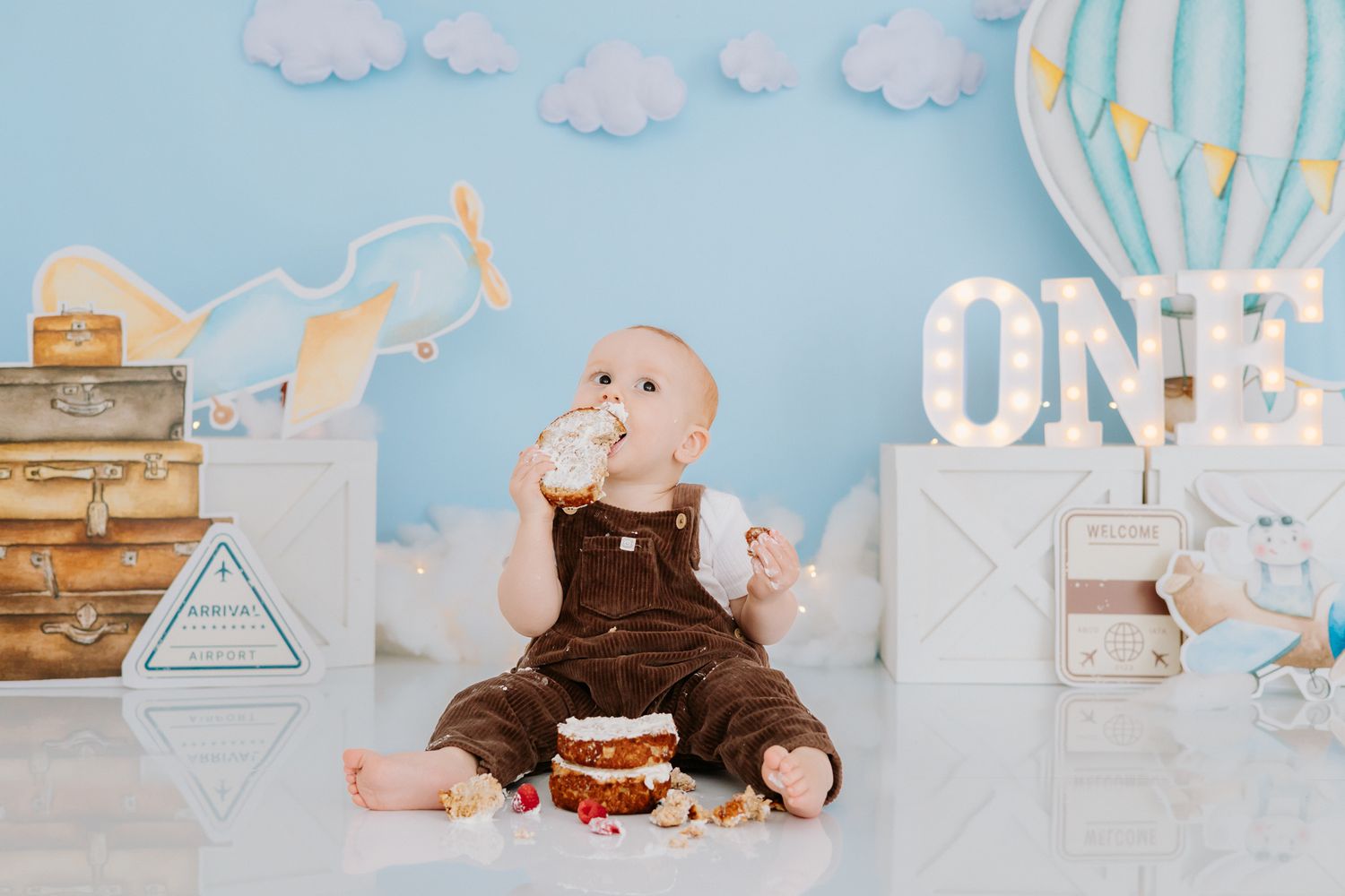 Baby in brown overalls celebrates first birthday with smashed cake, surrounded by travel-themed decor and ONE marquee sign.