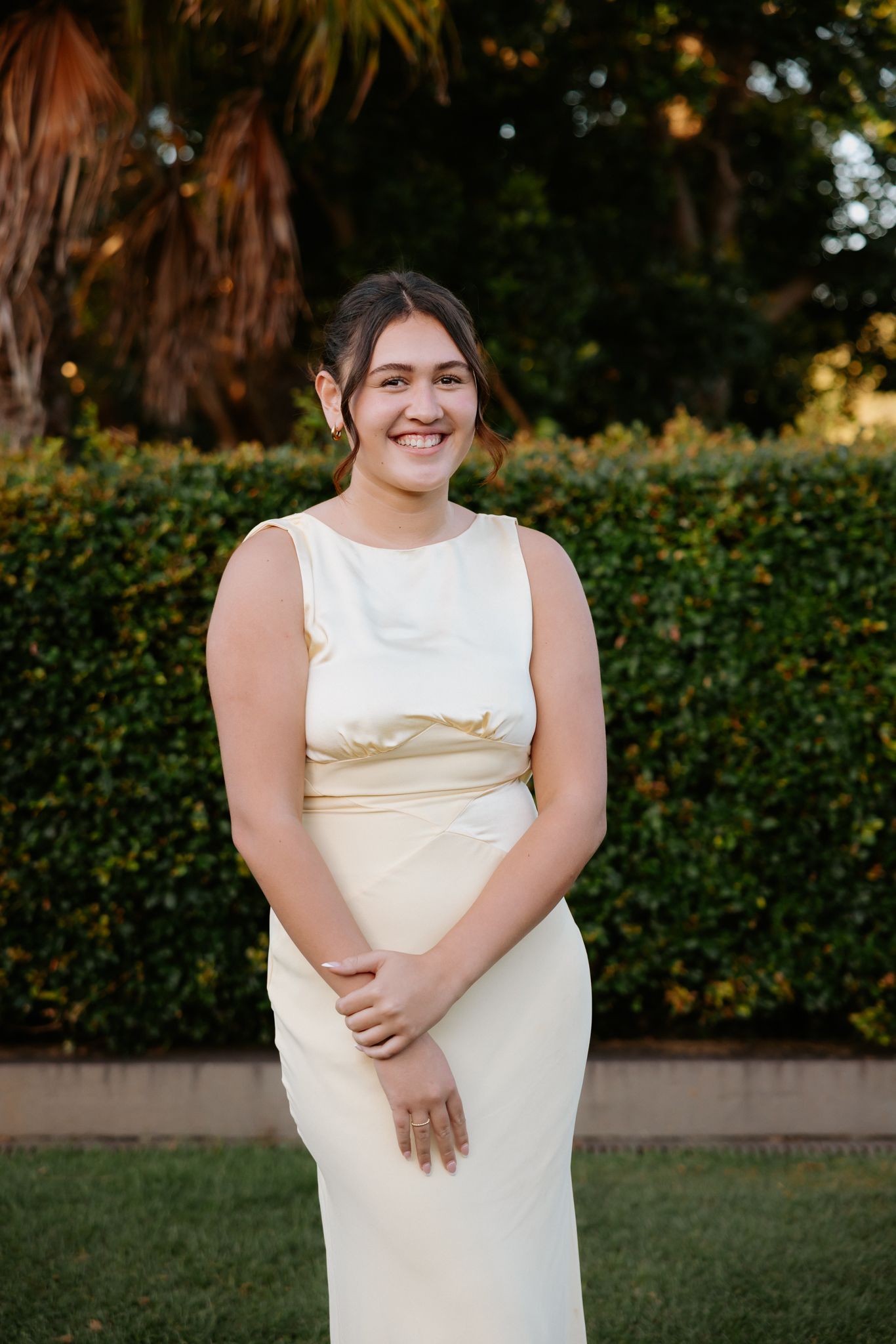 Young woman smiling in elegant cream sleeveless gown outdoors with green hedge background.