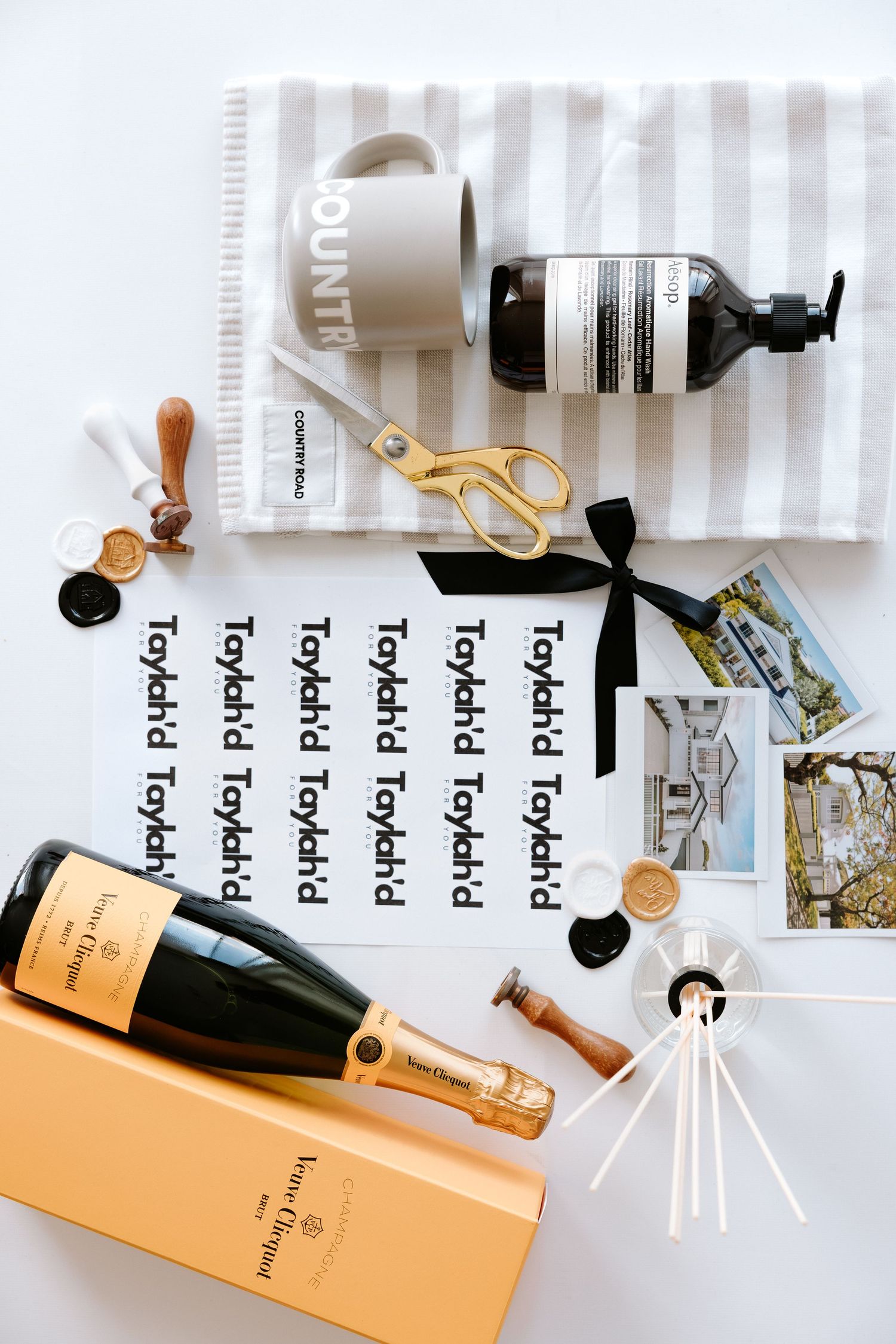 Flat lay of Veuve Clicquot champagne, Aesop soap, gold scissors, stickers, wax seals, and striped towel on white surface.