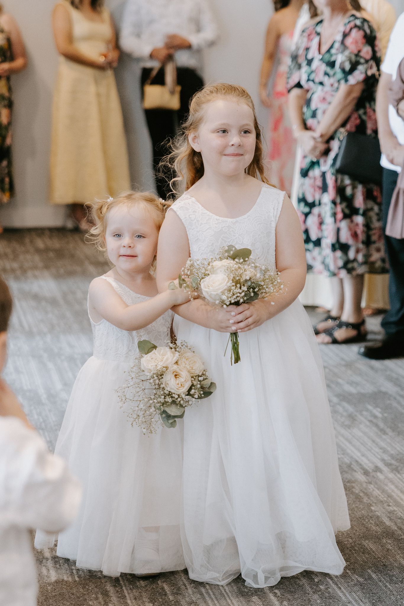 Two young flower girls in white dresses holding bouquets of white roses and baby's breath at a wedding ceremony.
