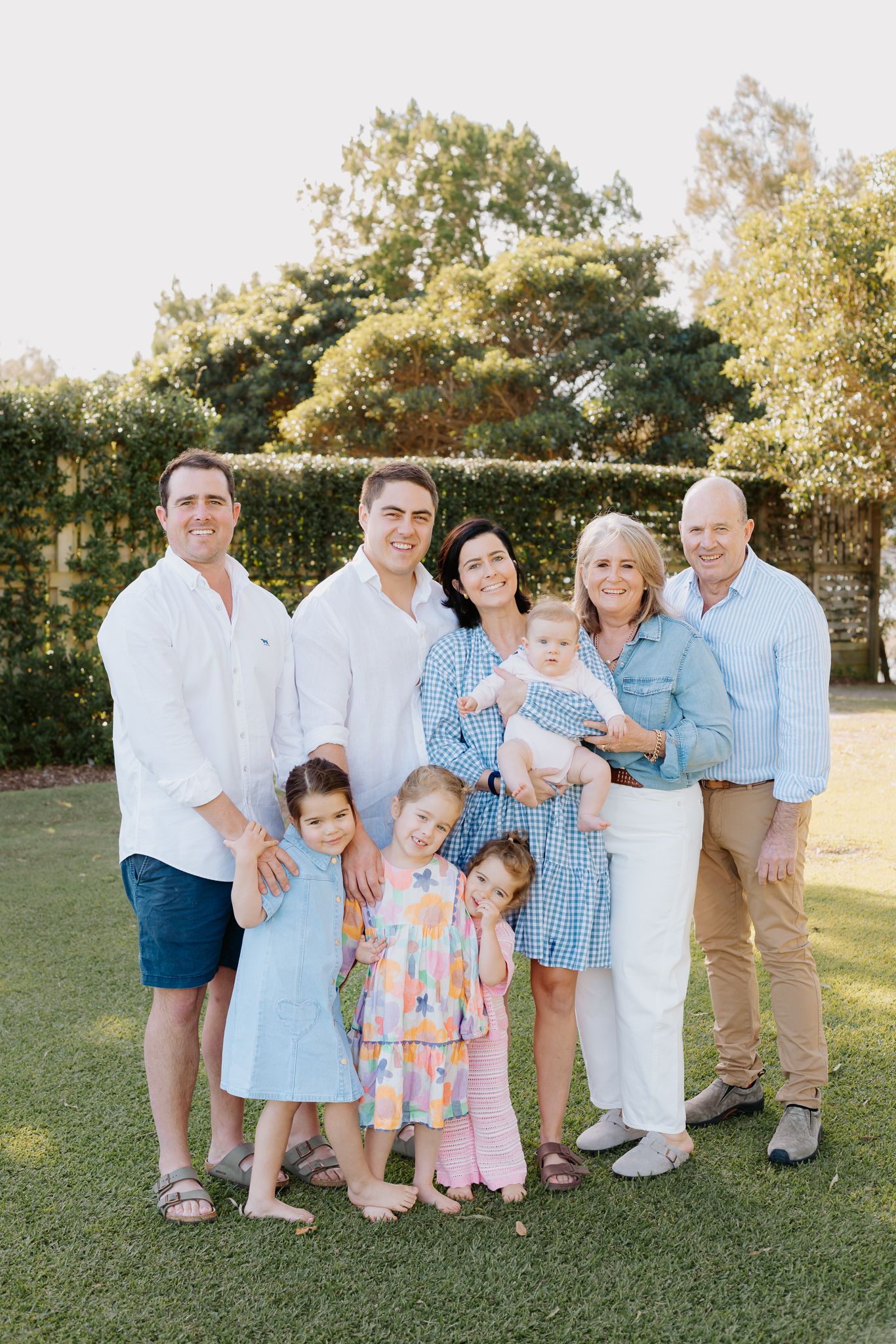 Multi-generational family portrait outdoors on green lawn, adults and young children smiling in coordinated blue and white outfits.