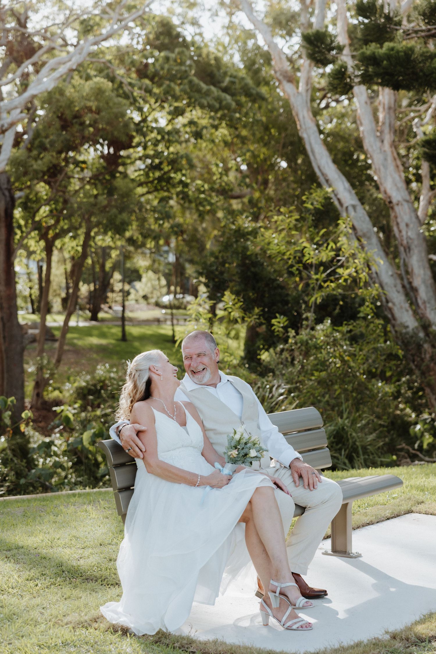 A couple in wedding attire sits together on a white bench in a lush garden setting with eucalyptus trees.