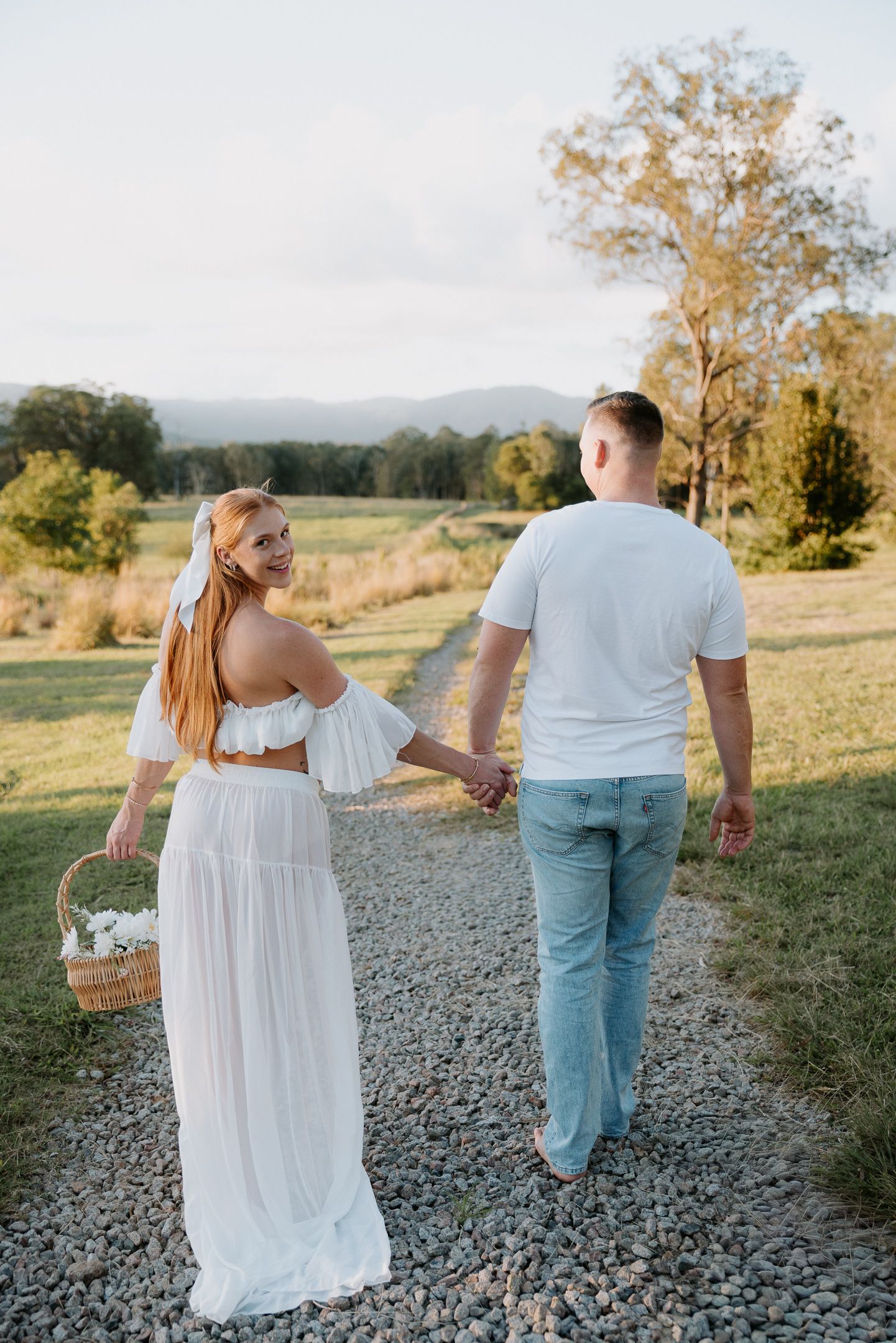 A couple walks hand-in-hand on a gravel path, woman in white dress holding flower basket, man in white tee and jeans, scenic countryside.