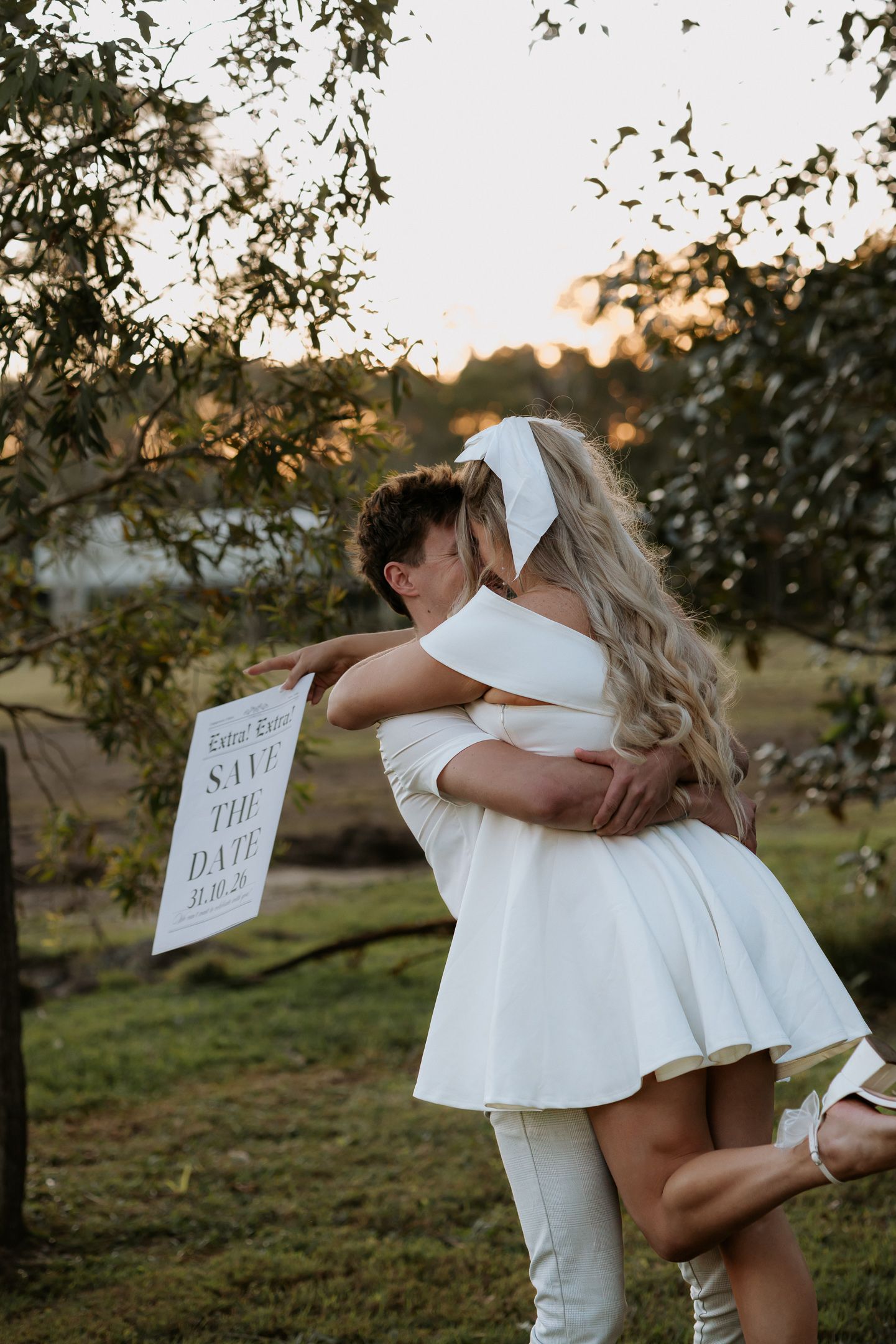 Couple in white outfits share a romantic embrace outdoors, holding a Save the Date sign at golden hour.