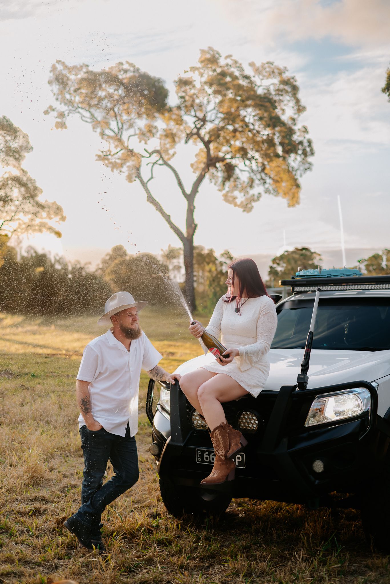 Couple celebrating with champagne beside a white 4WD at golden hour in a rural Australian field.