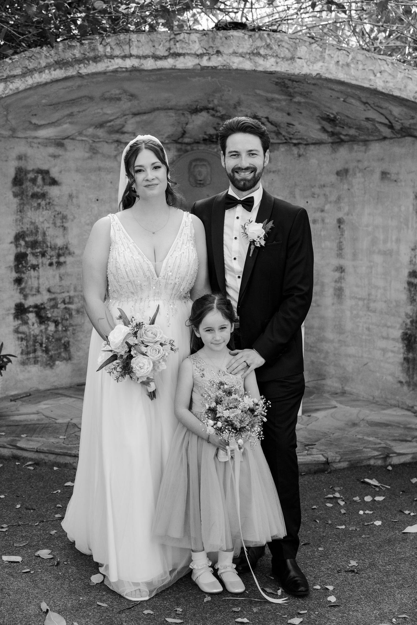 Black and white wedding photo of bride and groom with flower girl posing in front of stone wall.