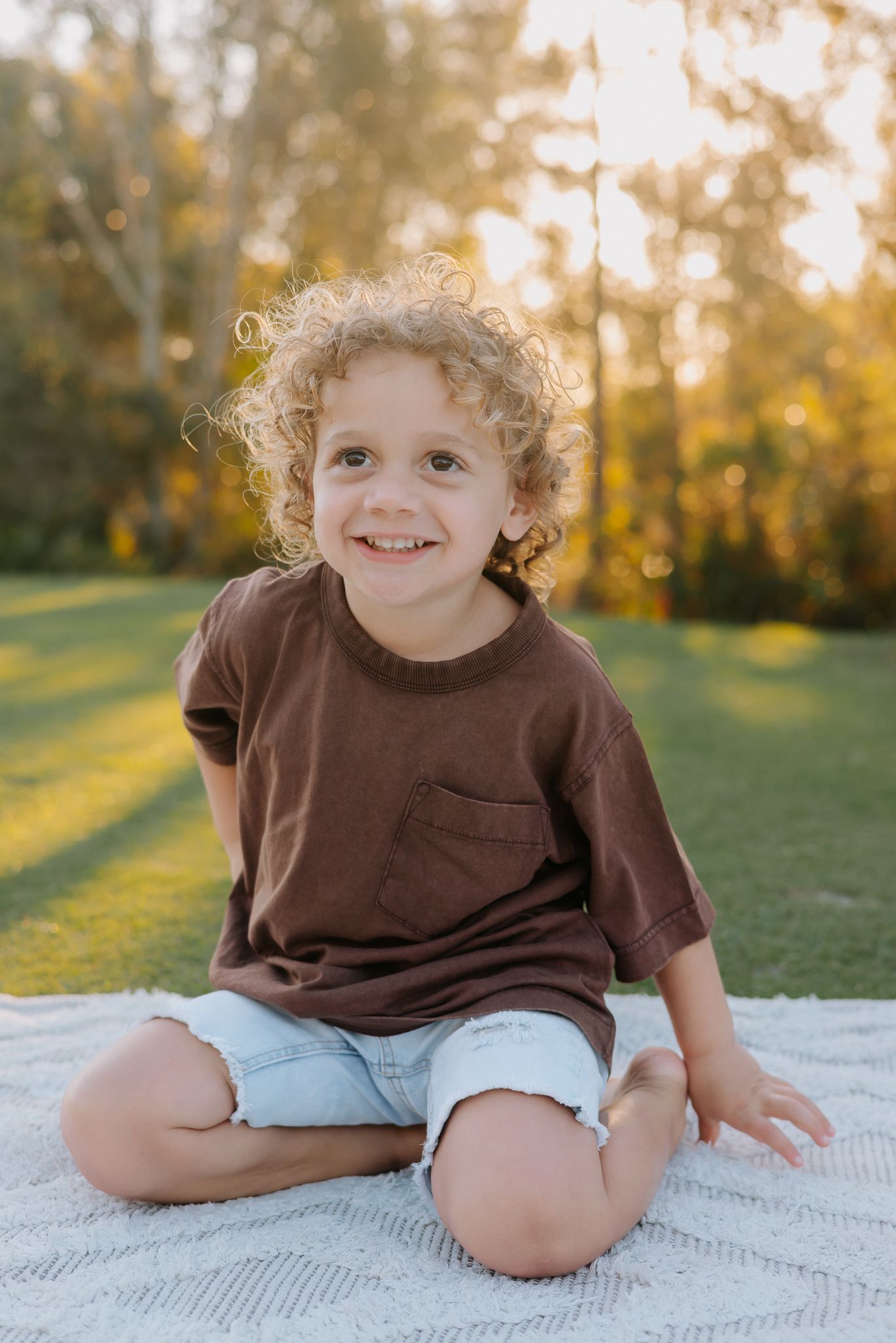 Smiling curly-haired boy sitting cross-legged on a blanket outdoors in golden sunlight wearing a brown t-shirt and denim shorts.