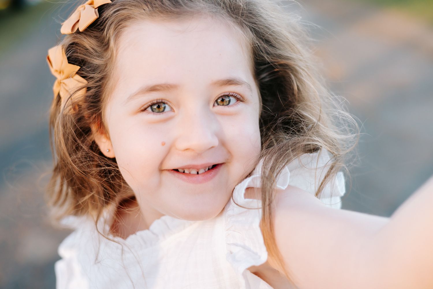 Smiling young girl with curly hair and floral hair clip wearing a white dress outdoors.