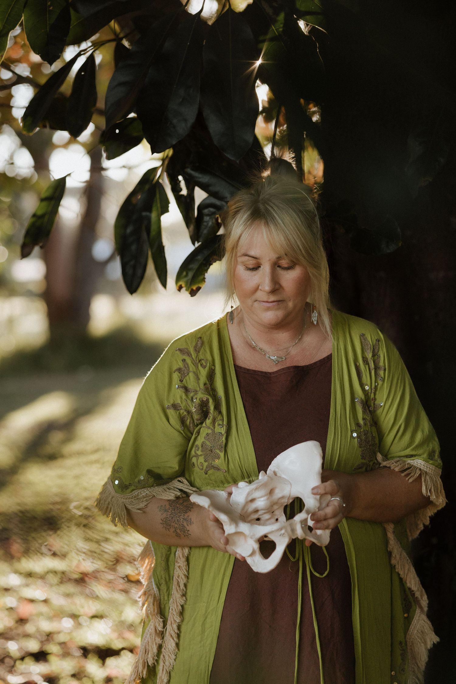 Blonde woman in green kimono holding a white pelvis bone model outdoors under a tree in natural light.