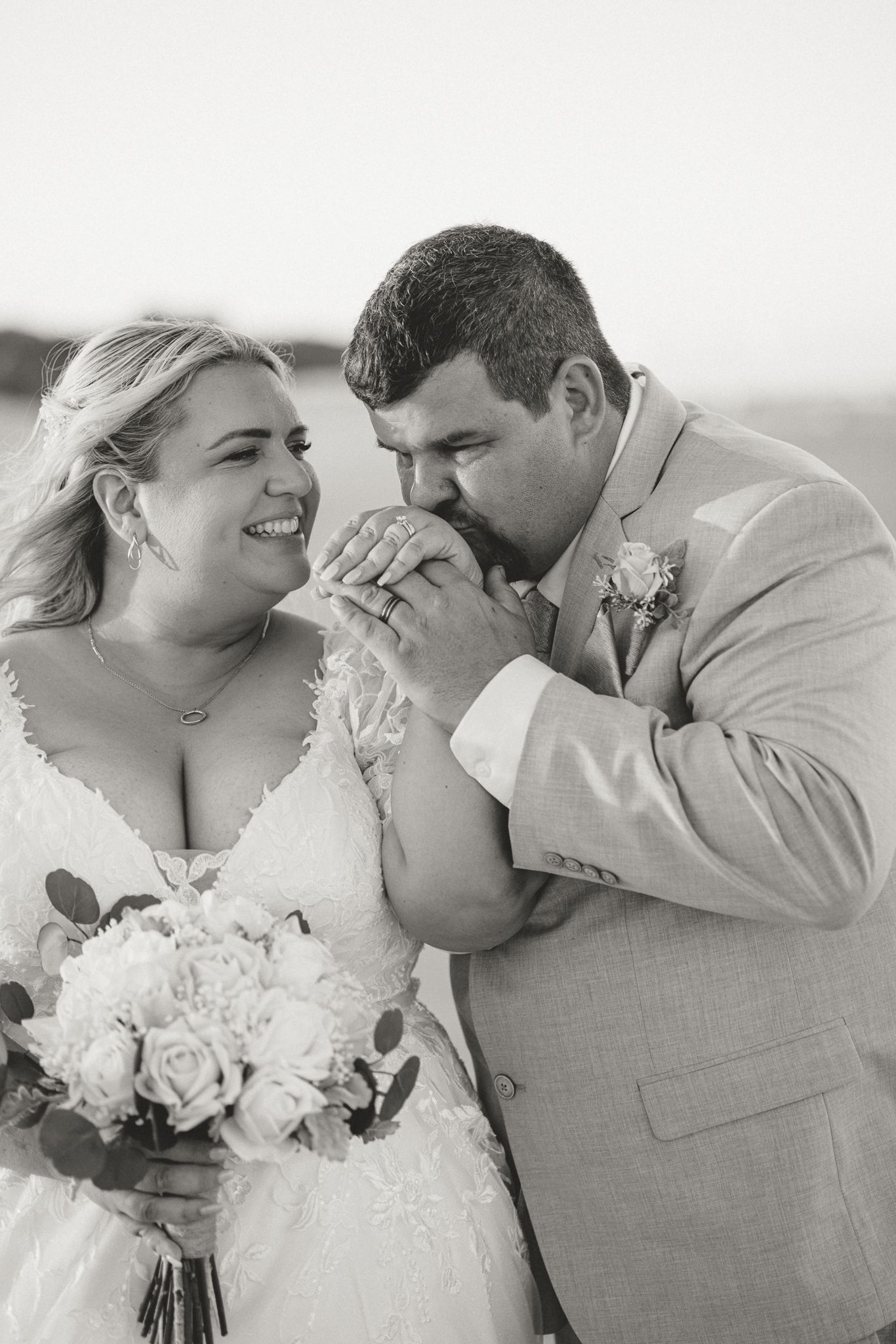Black and white photo of a groom kissing his bride's hand while she smiles, holding a white rose bouquet on their wedding day.