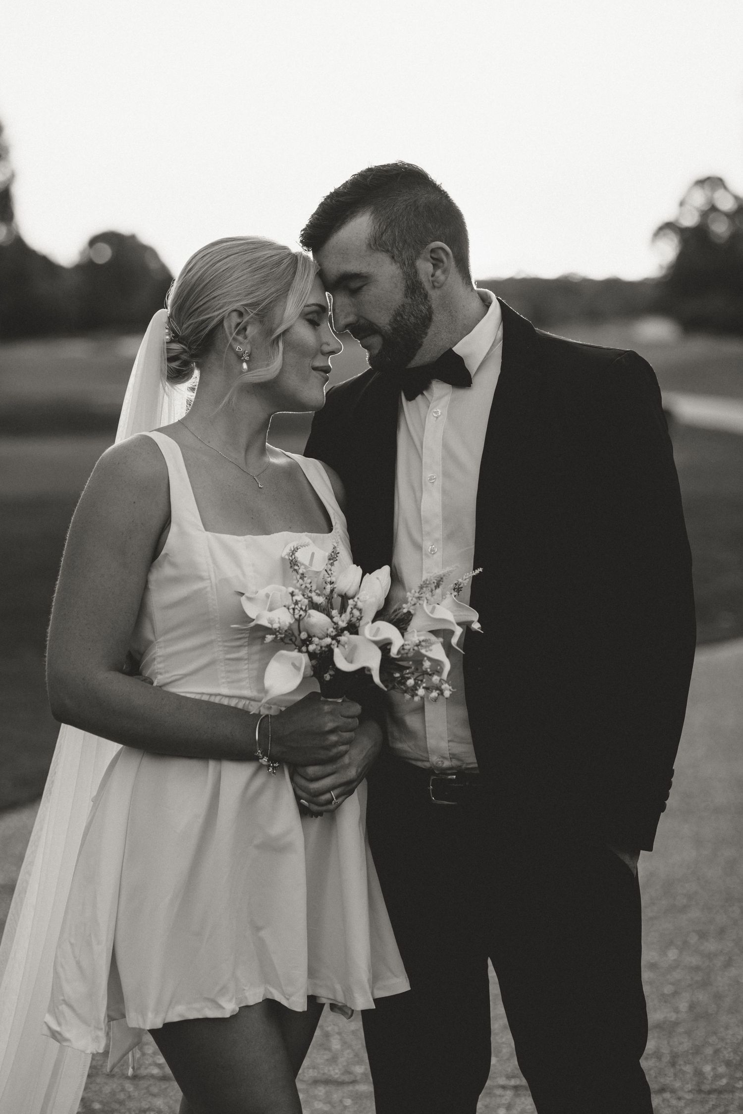 Black and white photo of a bride in short dress holding bouquet and groom in tuxedo sharing an intimate moment outdoors.