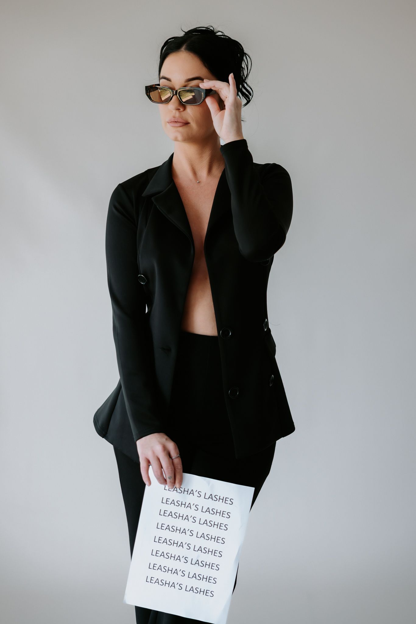 Woman in black blazer holding Leasha's Lashes sign, adjusting sunglasses against white background.