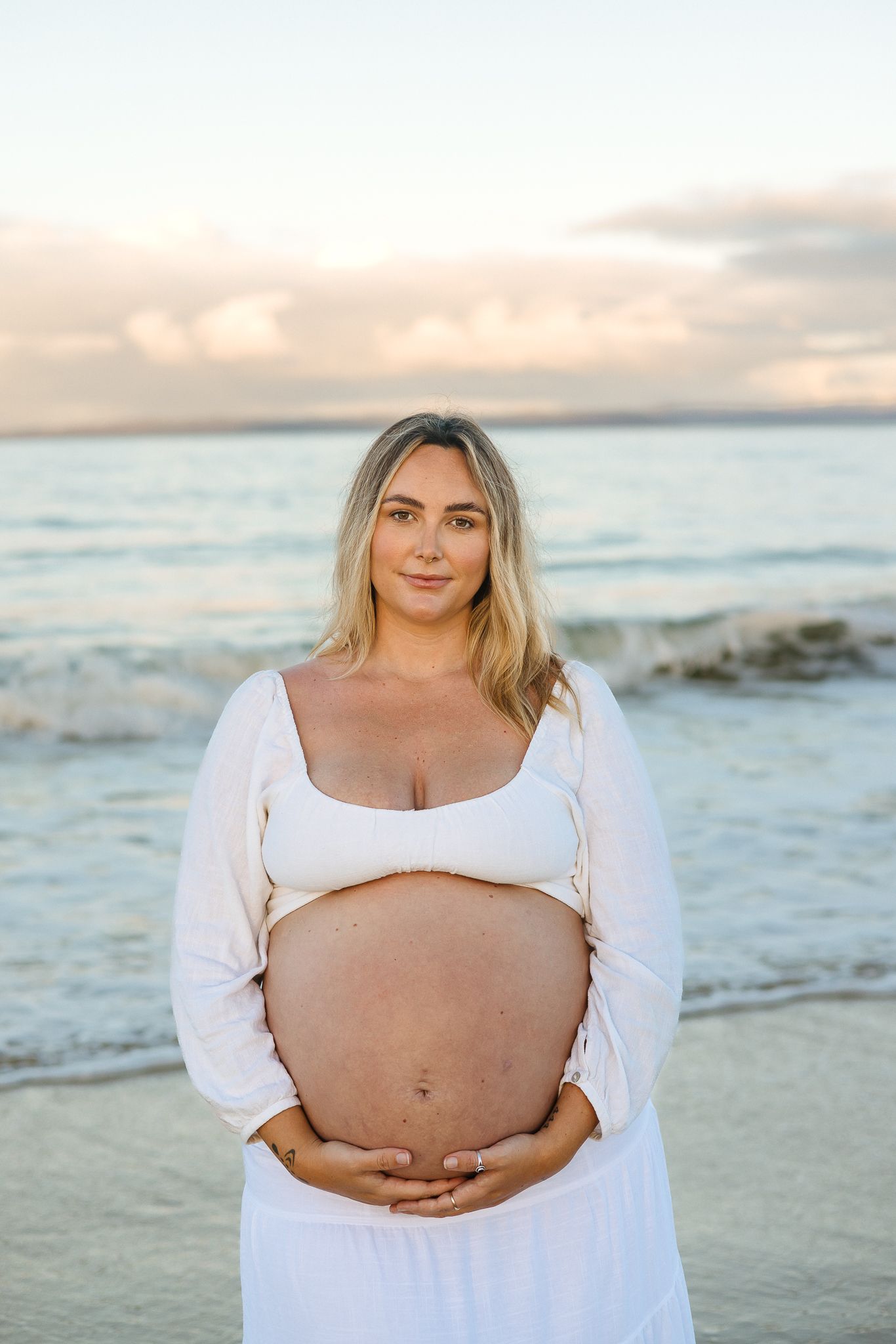 Pregnant woman in white dress holding baby bump on beach at sunset.