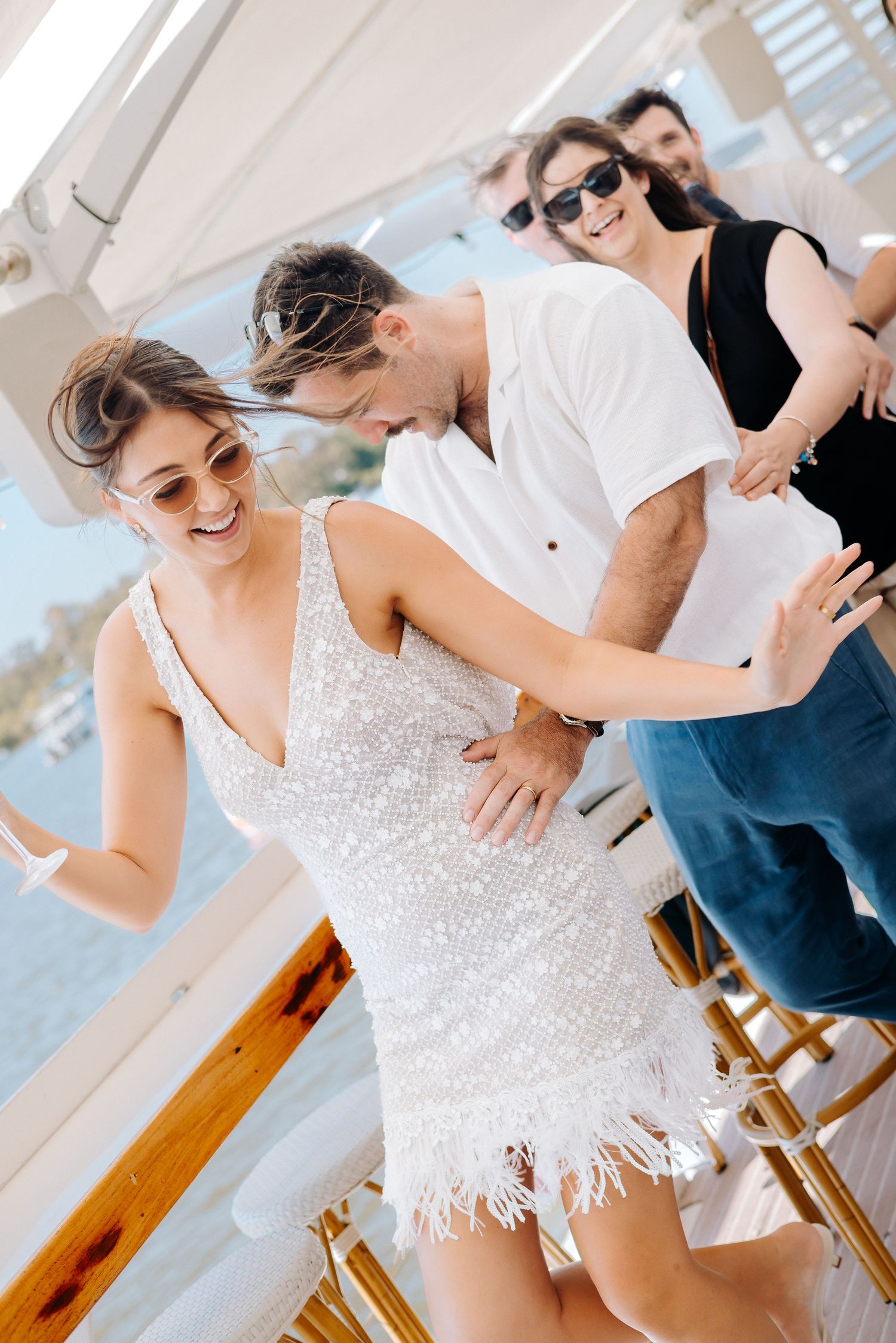 Couple dancing joyfully on a yacht deck, woman in white sequin dress, friends celebrating in background.