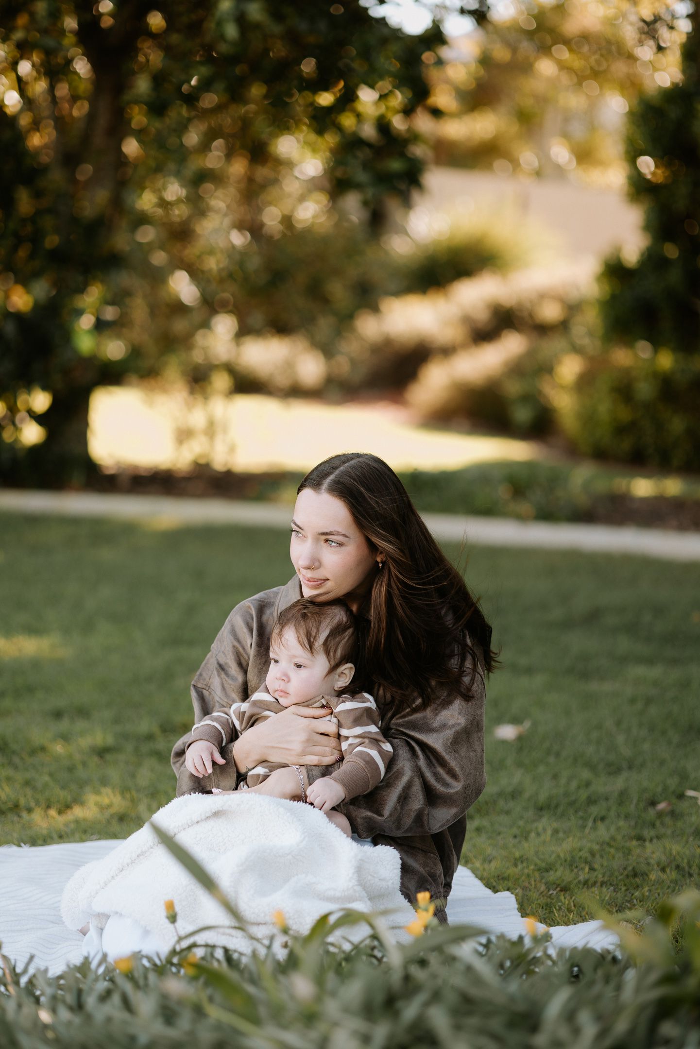A parent and baby sit together on a white blanket in a sunny park during autumn.