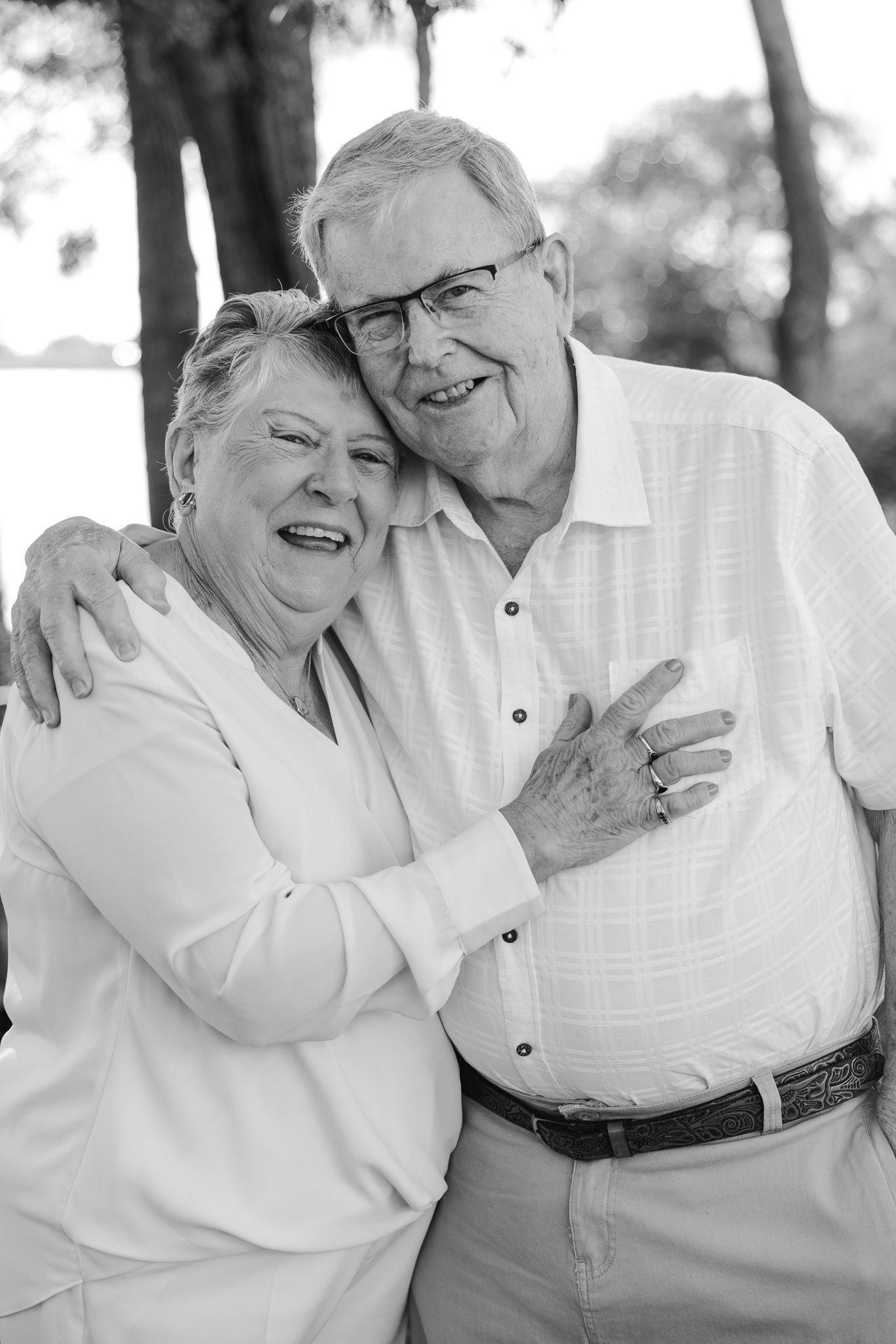 Black and white portrait of a happy elderly couple embracing outdoors, smiling warmly near trees.
