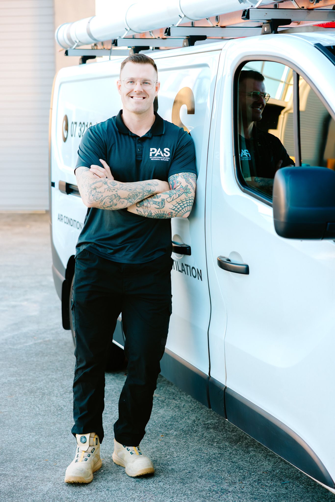 Smiling tradesman in PAS uniform stands arms crossed beside a white work van with ladder rack.