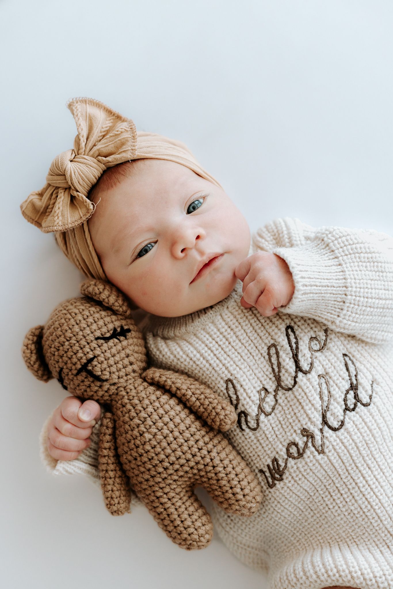 Newborn baby in a cream Hello World sweater with a tan bow headband, cuddling a brown crocheted teddy bear.