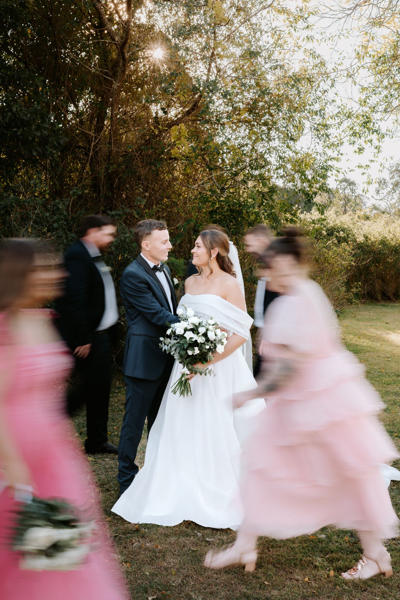 Bride and groom share a tender moment outdoors as bridesmaids in pink dresses walk past in a blur.