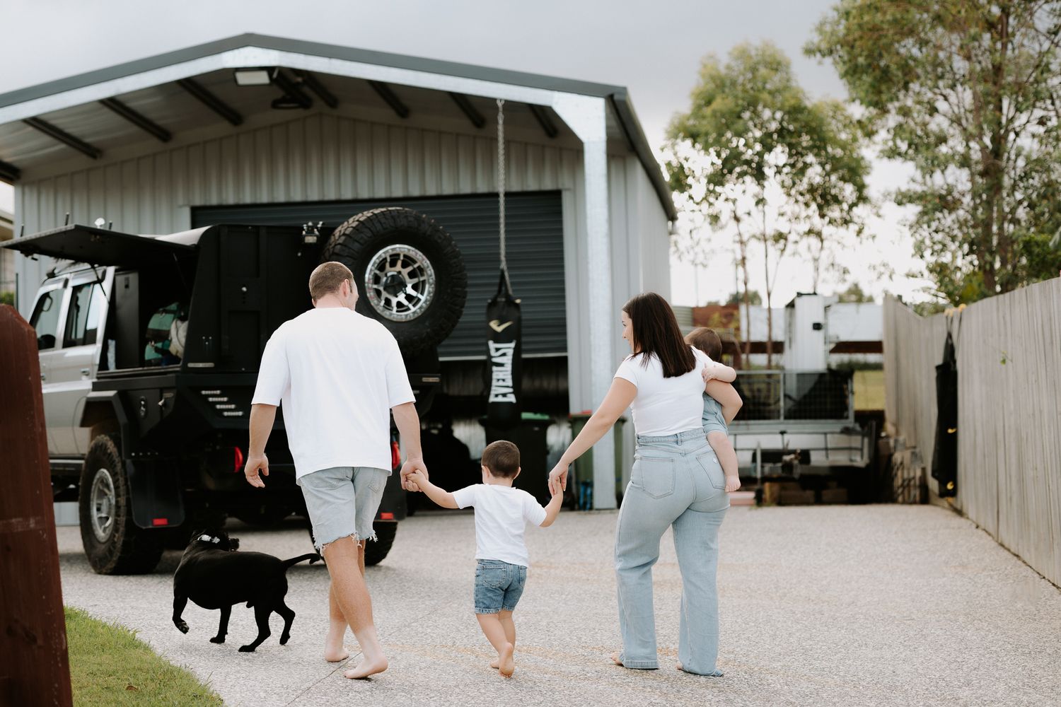 Family with toddler and black dog walking toward a 4WD garage on a gravel driveway.