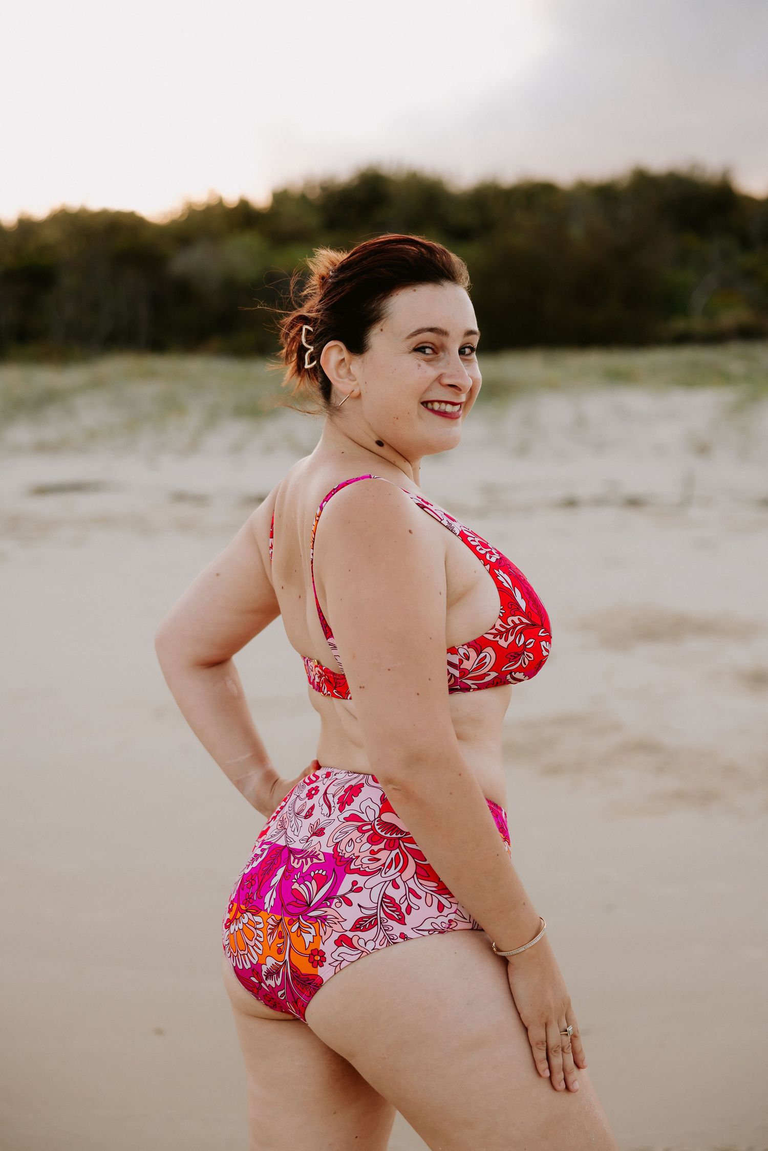 Woman in pink floral bikini posing on sandy beach at sunset with trees in background.