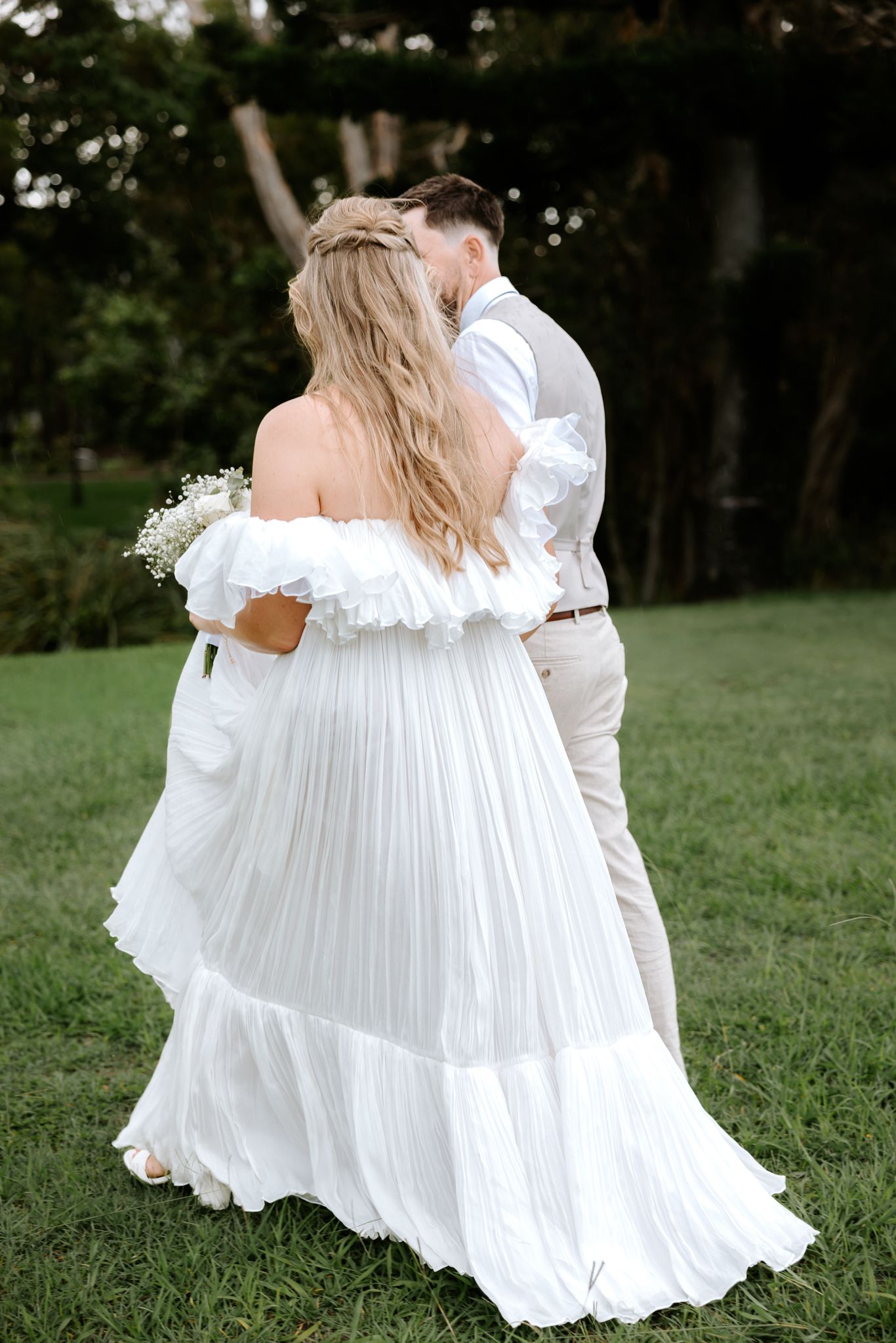 Bride in flowing off-shoulder white pleated gown and groom in beige vest walking together on green lawn.