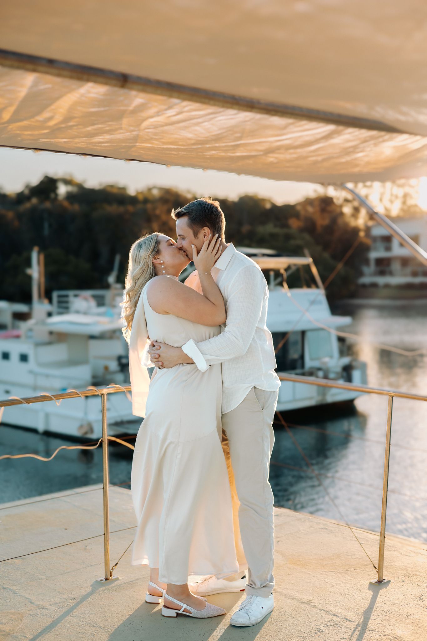 Couple kissing on a yacht deck at golden hour, dressed in elegant white outfits with marina boats in background.