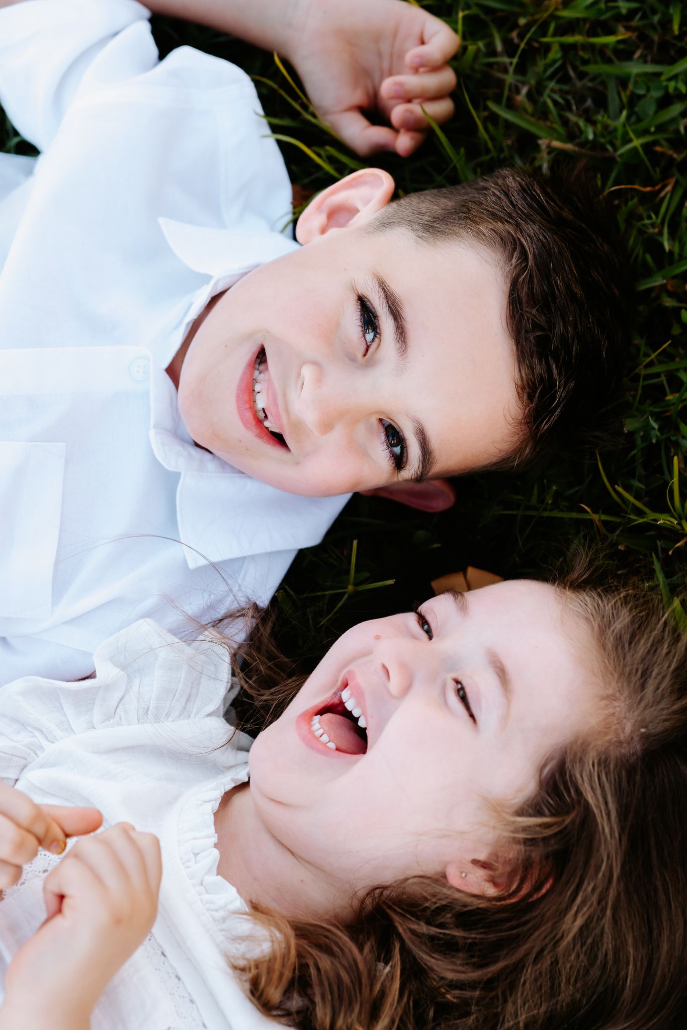 Two happy children in white outfits lay on green grass laughing and smiling together outdoors.