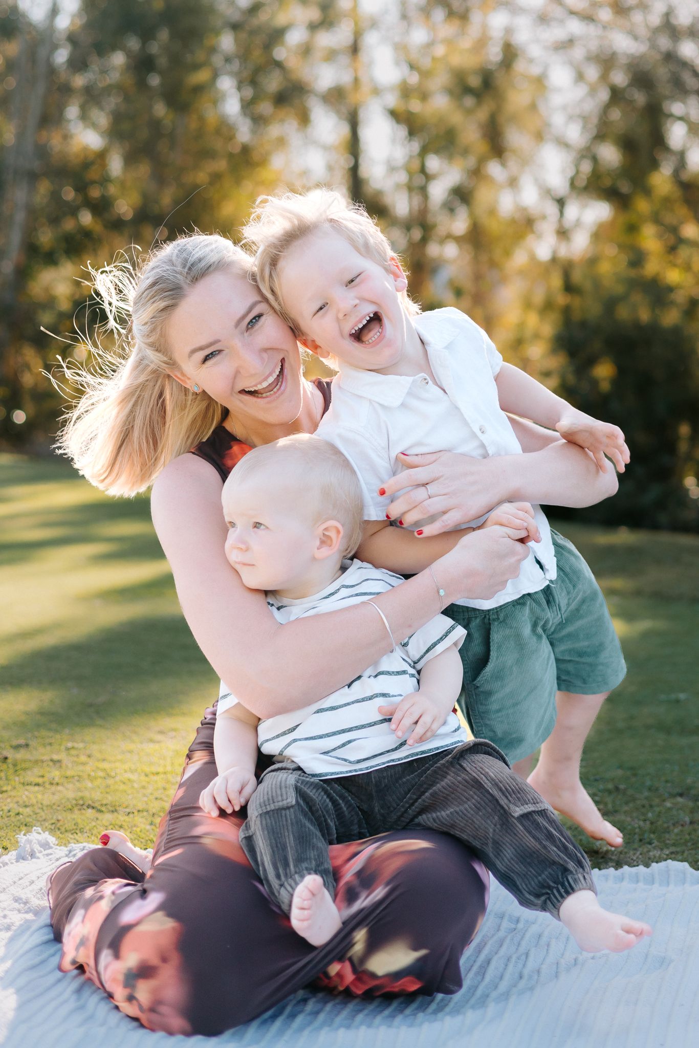A joyful mother sits on a blanket outdoors, laughing with her two young children on a sunny day.