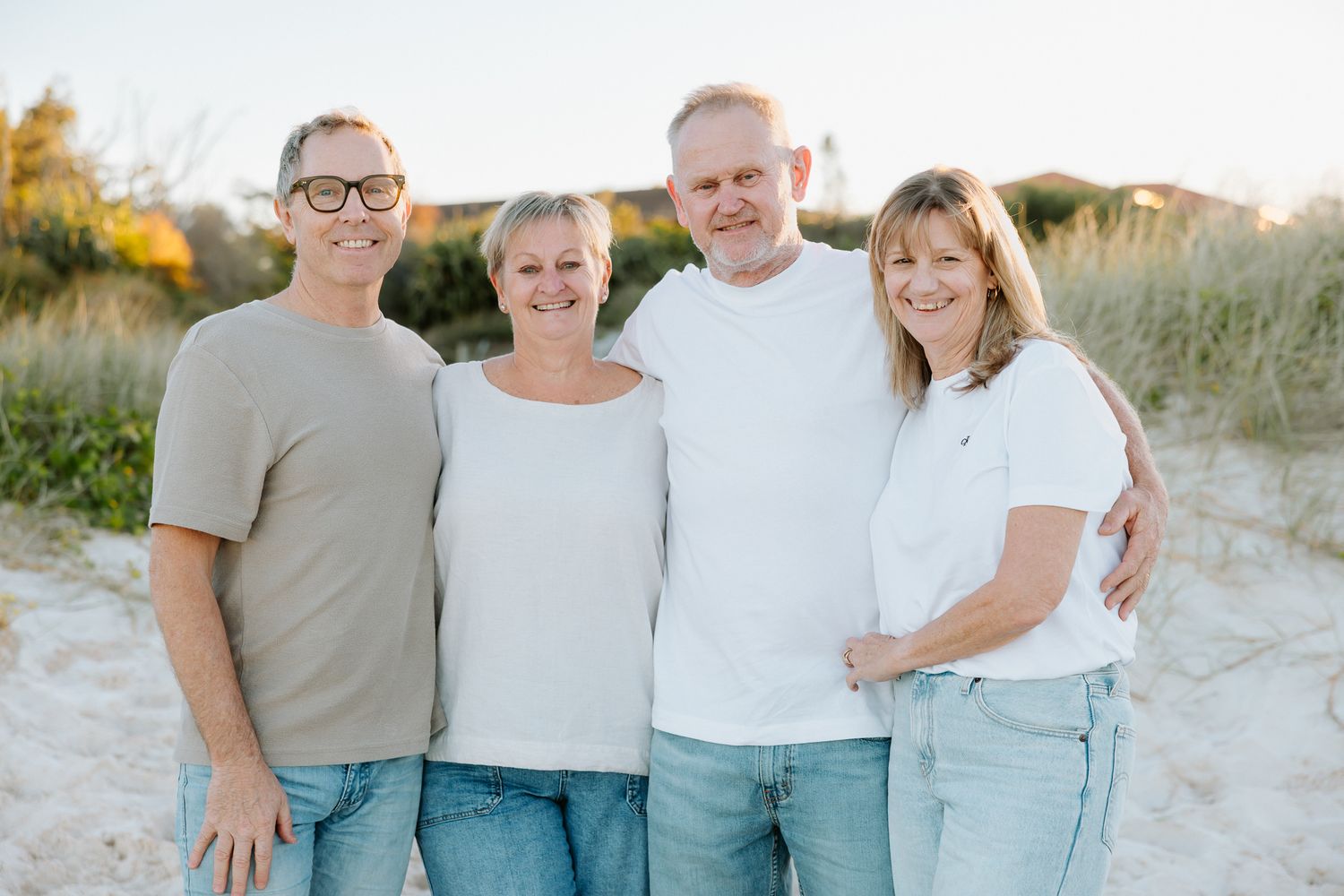 Four adults smiling together on a sandy beach, wearing casual white and beige tops on a sunny day.