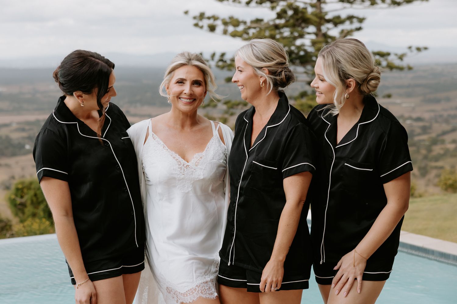 Bride in white with three bridesmaids in black pajamas smiling by a pool with scenic views.