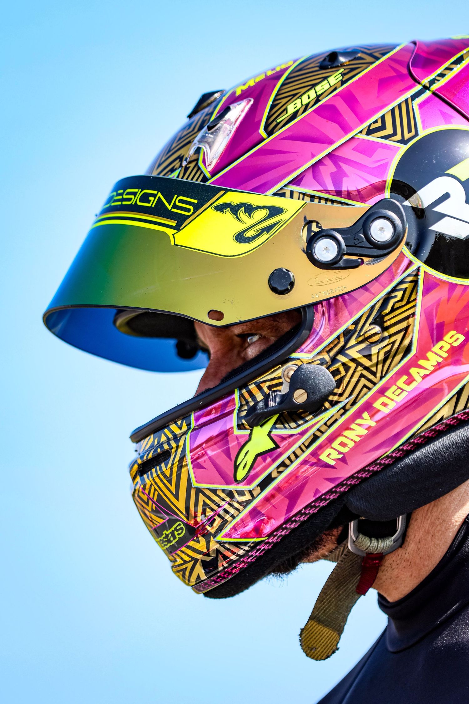 Colorful pink and yellow racing helmet with iridescent visor worn by driver Ronny Dielamps against blue sky.