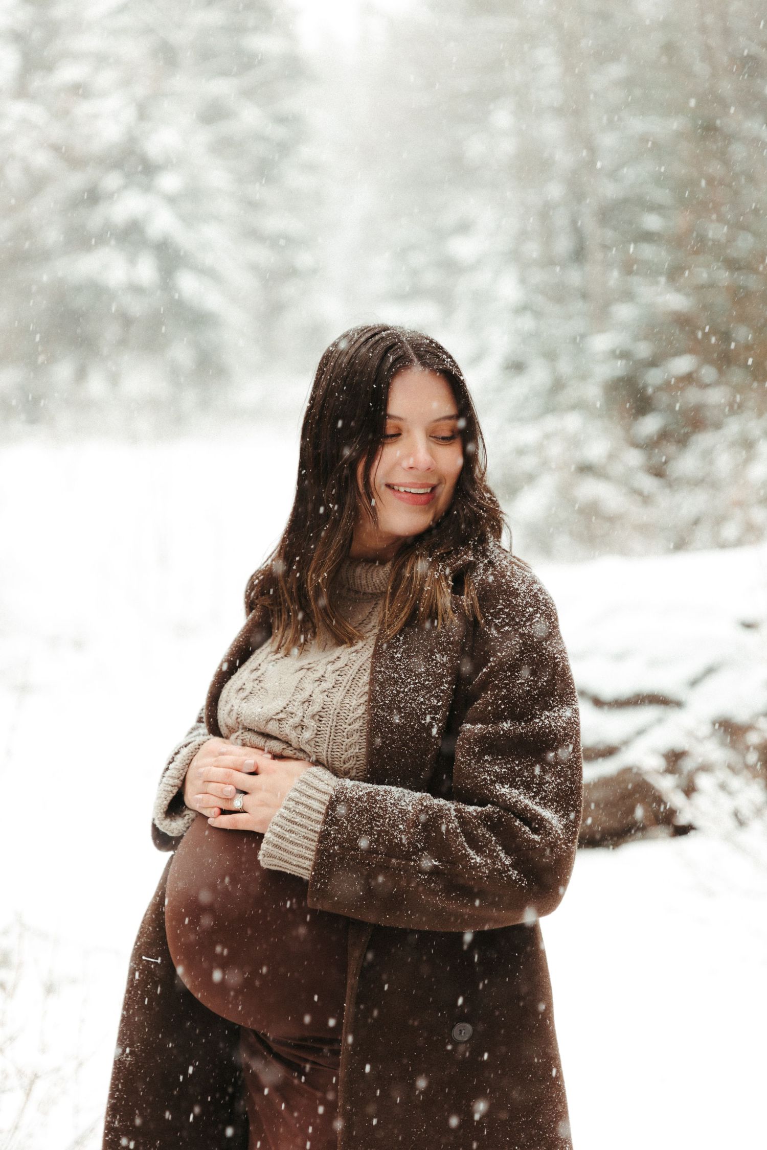 A person in a brown sweater smiles while standing in falling snow.