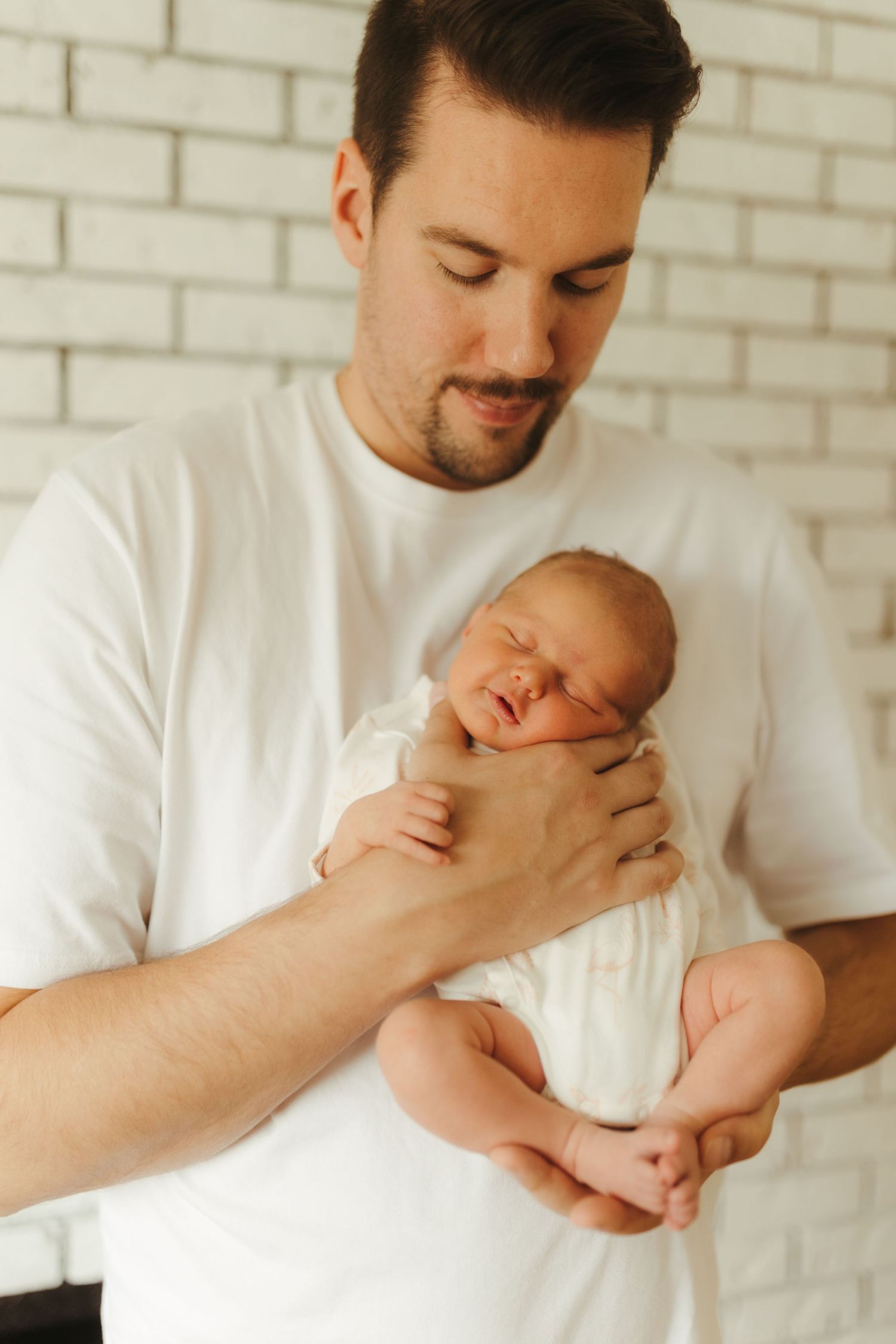 A tender moment between parent and sleeping newborn baby against a white brick wall background.