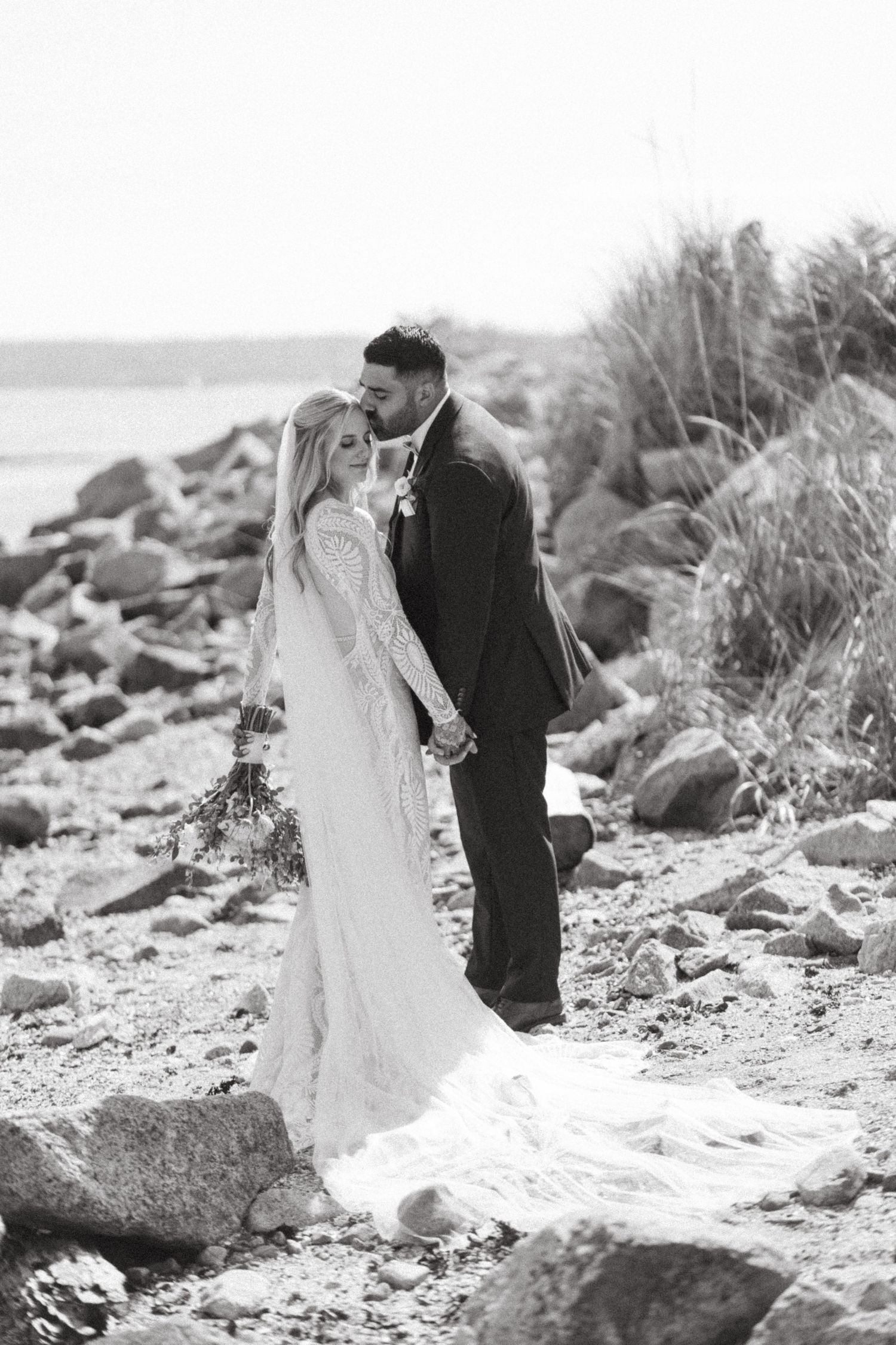 A romantic couple in wedding attire shares intimate moments along a rocky beach shoreline in black and white photography in Vancouver