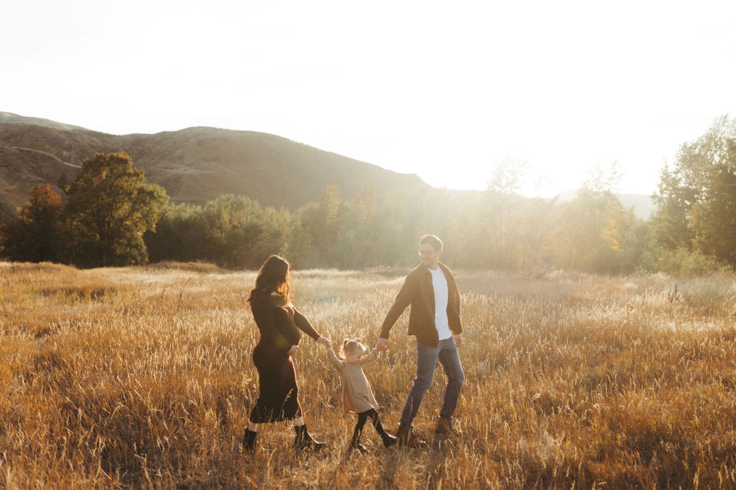A couple and child walk through a golden field at sunset with mountains in the background.