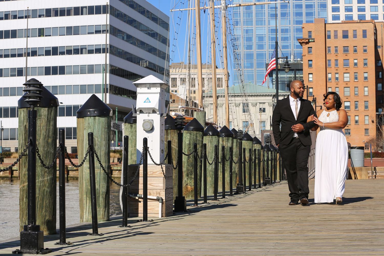 People stand along a wooden pier railing overlooking the water with city buildings visible in the background.