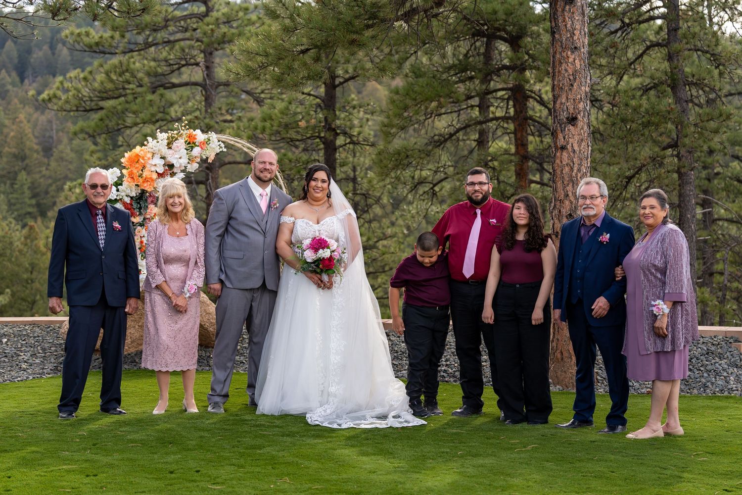 Wedding party and guests gathered for group photos on a grassy lawn surrounded by pine trees.