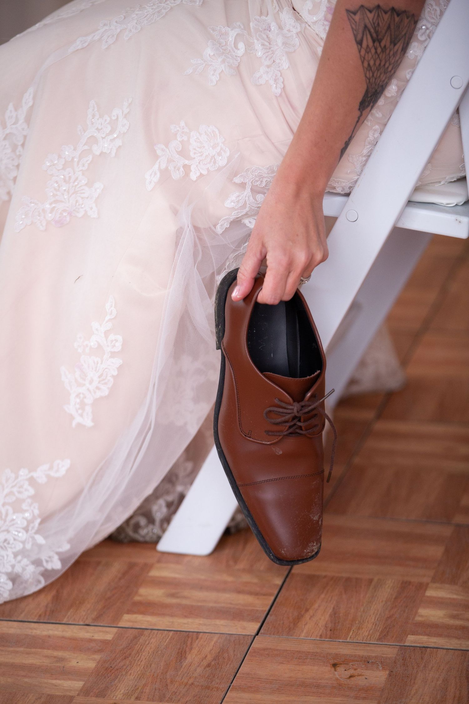 Close-up of white lace wedding dress and brown leather boots on hardwood floor.