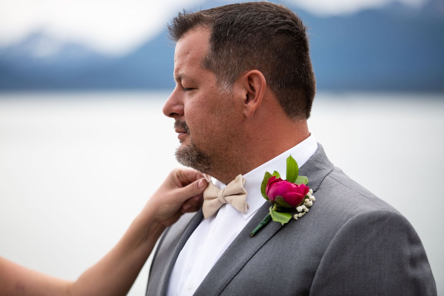 Someone adjusts a bowtie on a groom wearing a gray suit with pink boutonniere against a mountain backdrop.