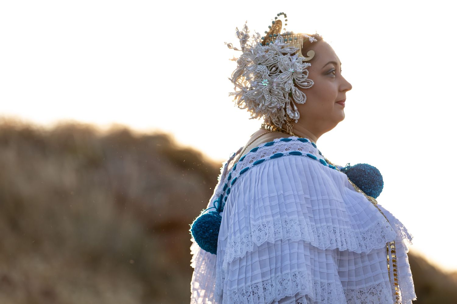 Dancer with ornate headpiece performs graceful movements in white costume against rolling hills.