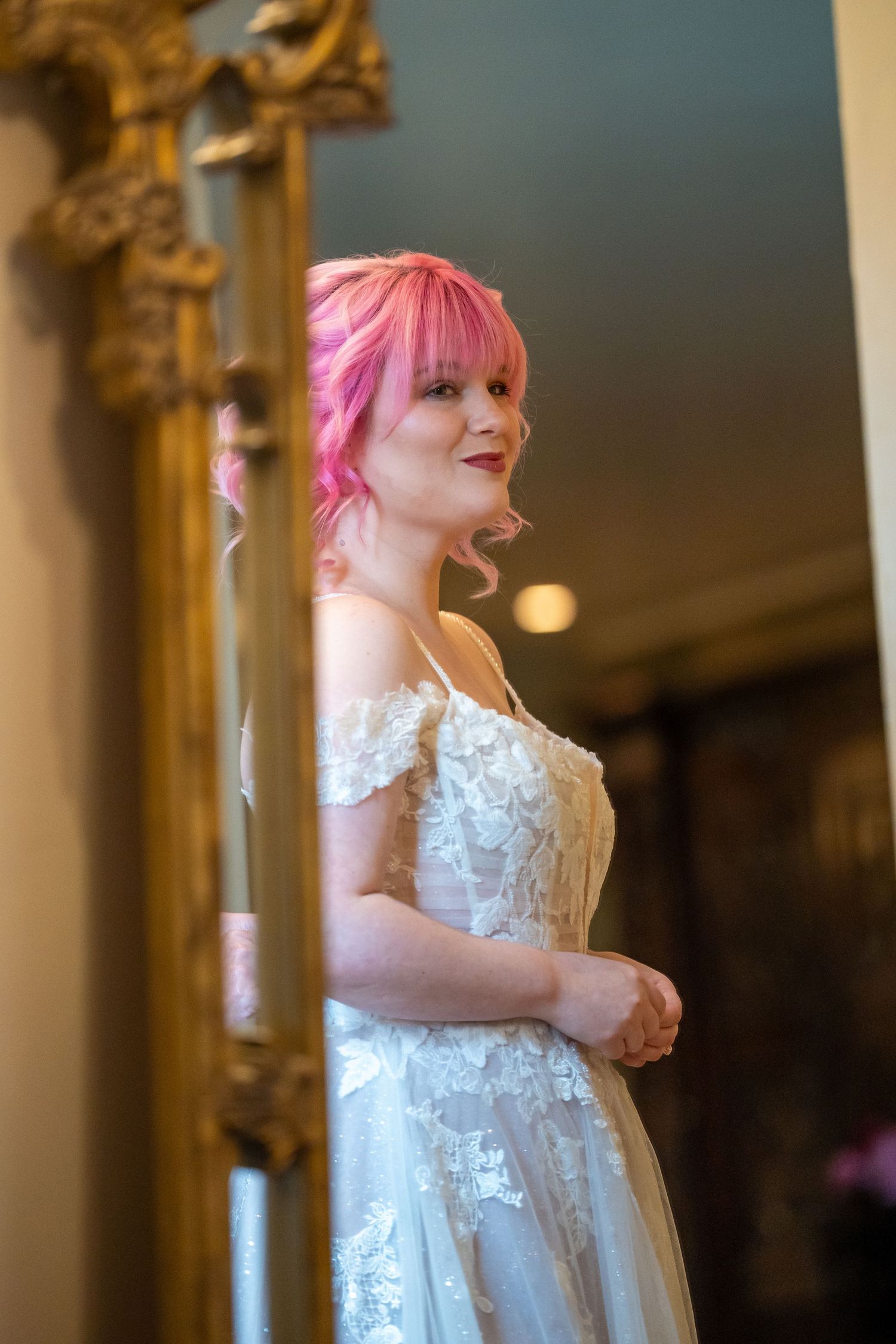 Bride in lace wedding gown poses elegantly beside ornate gold mirror.