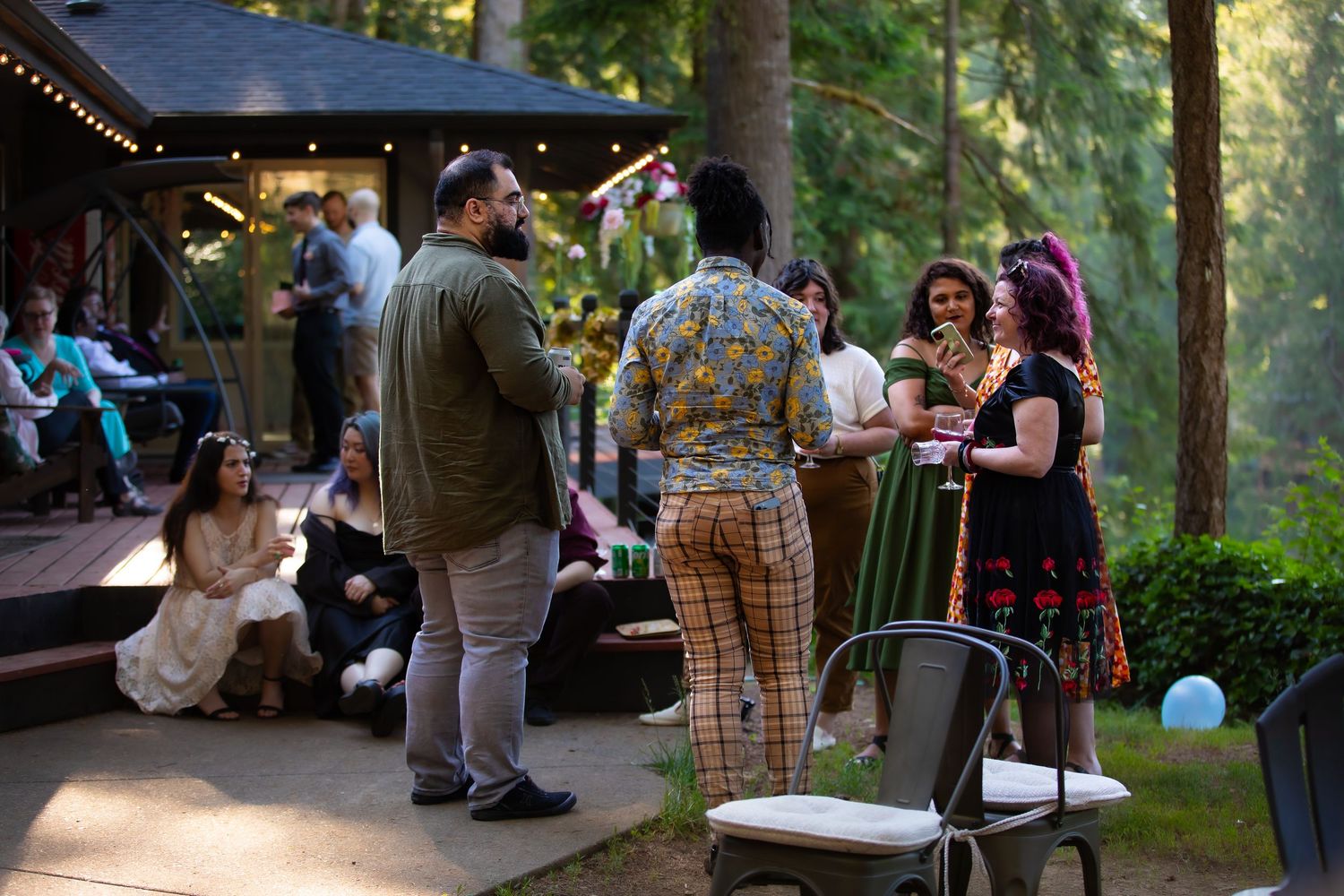 String lights illuminate a wooded outdoor gathering space with guests mingling.