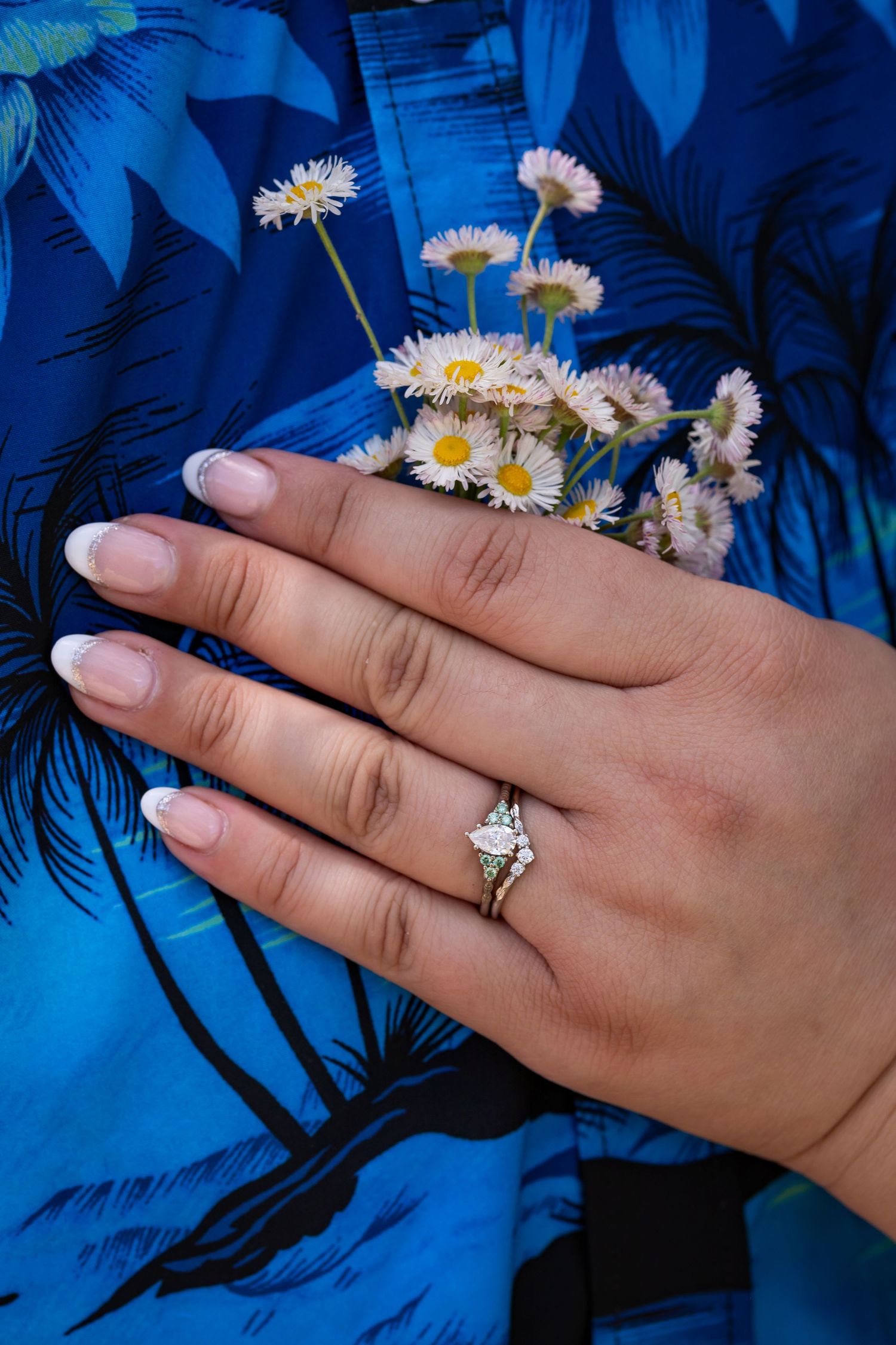 A hand adorned with an engagement ring holds delicate white daisies against a blue floral background.