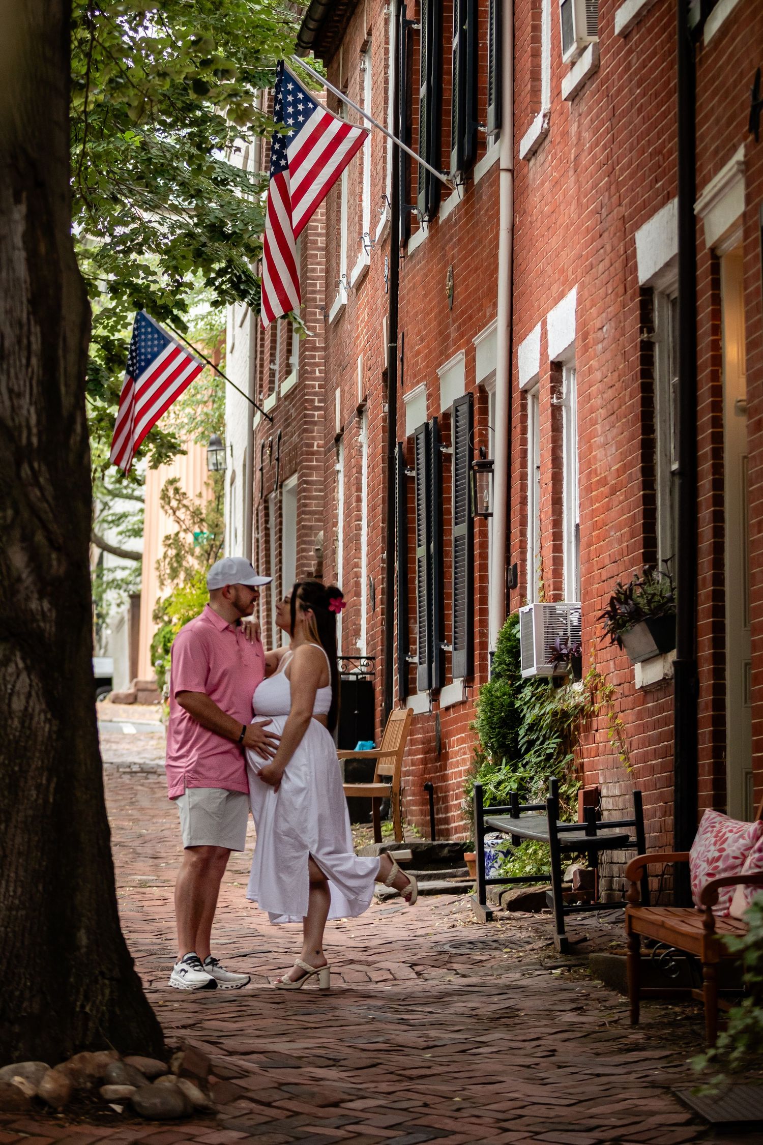 A couple shares a romantic moment on a historic cobblestone street lined with brick buildings and American flags.