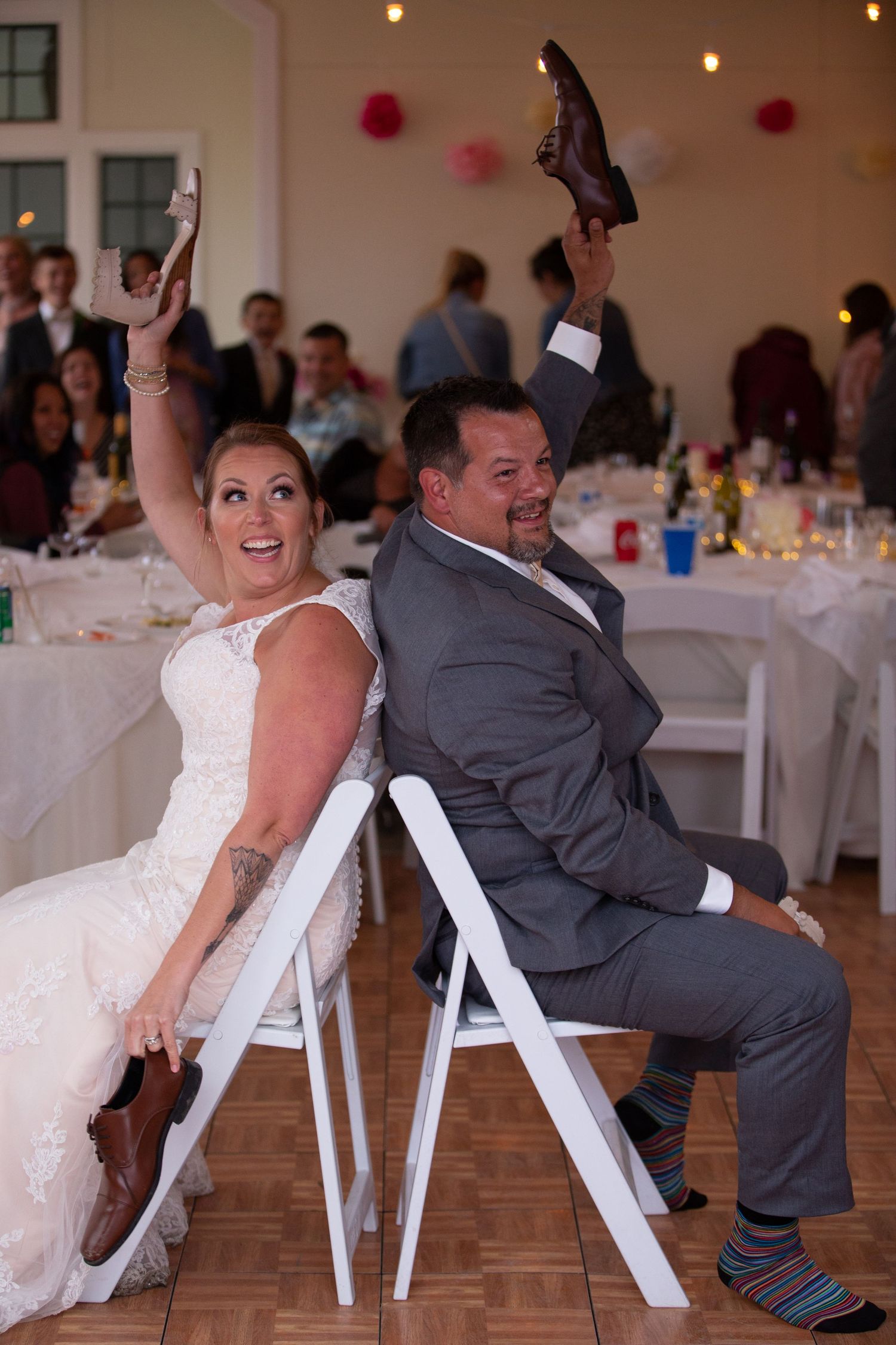 Newlyweds playing shoe game at wedding reception while seated on white chairs.